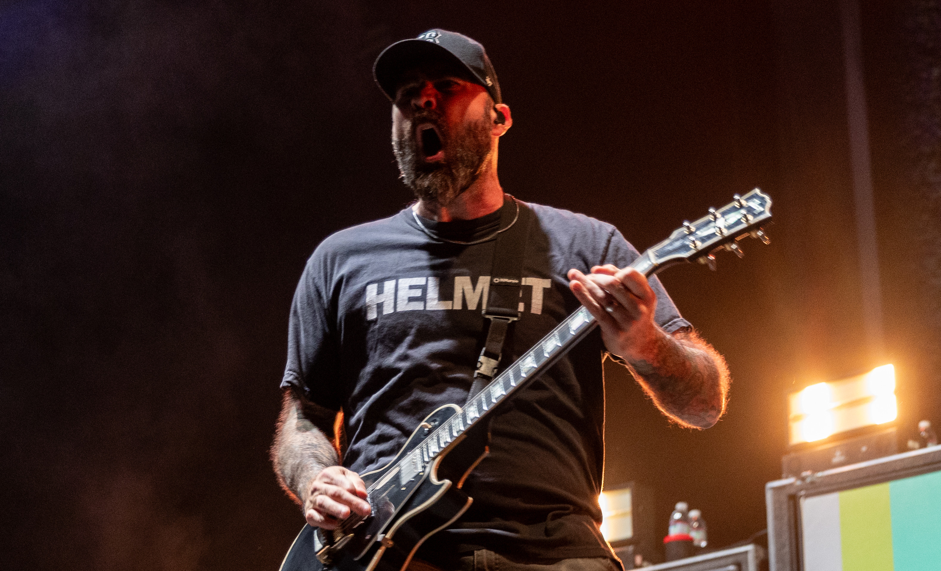 Chad Gilbert wearing a "Helmet" shirt and cap plays energetically on stage, with a colorful background and a musician at a DJ booth.