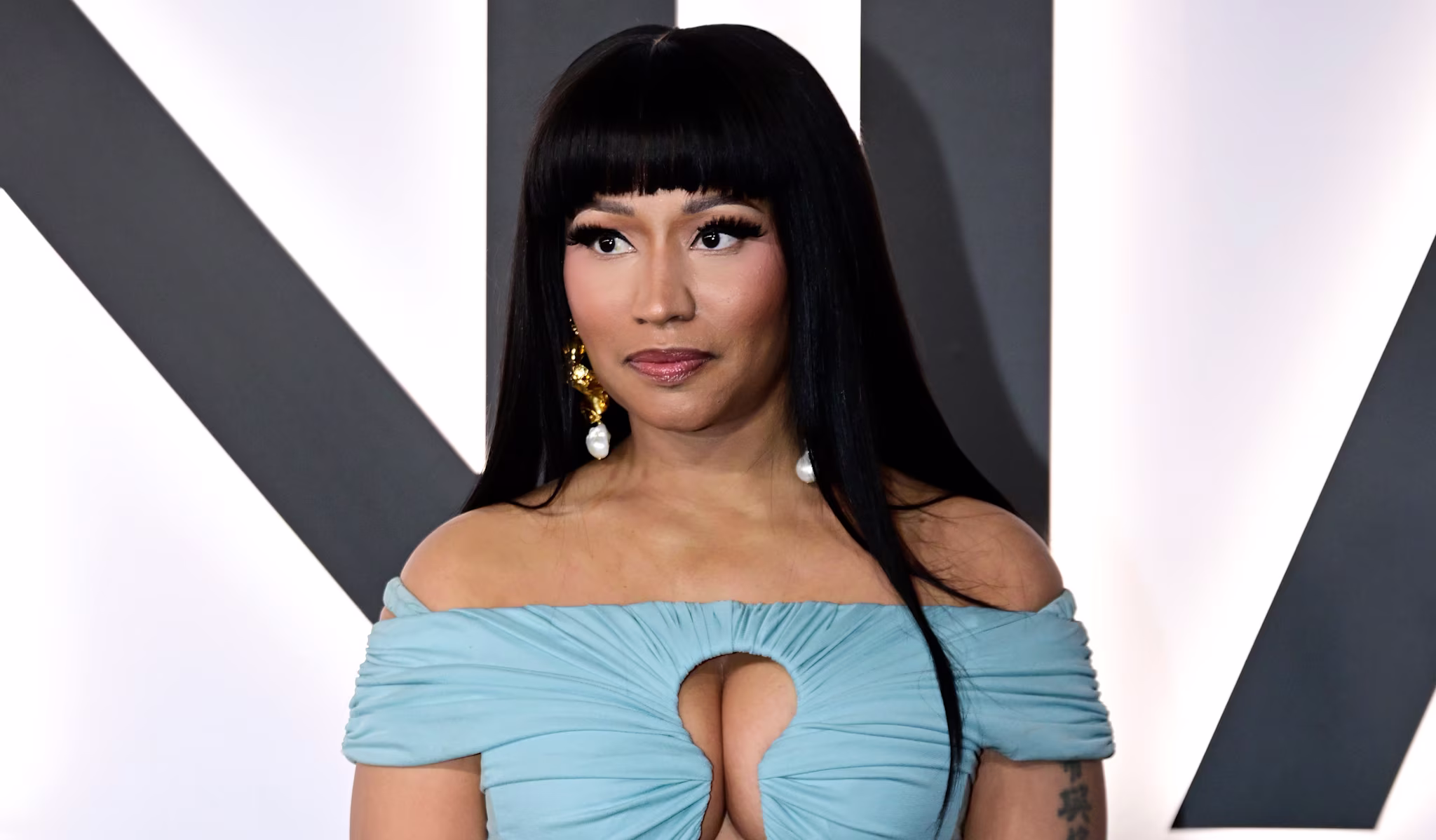 Nicki Minaj in a light blue off-shoulder dress with gold bracelets, standing against a neutral background.