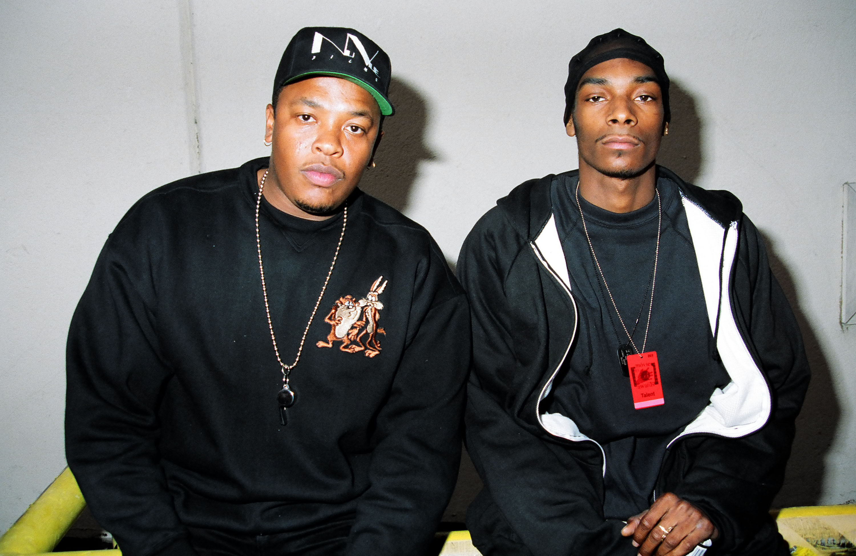 Dr. Dre and Snoop Dogg sitting together, both wearing black outfits. Dr. Dre has a cap, and Snoop Dogg wears a beanie.