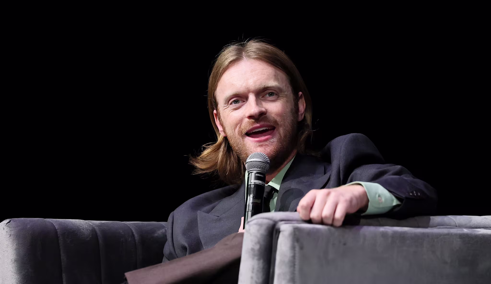 Finneas O'Connell speaking into a microphone while seated on a grey couch, wearing a dark jacket and light shirt.
