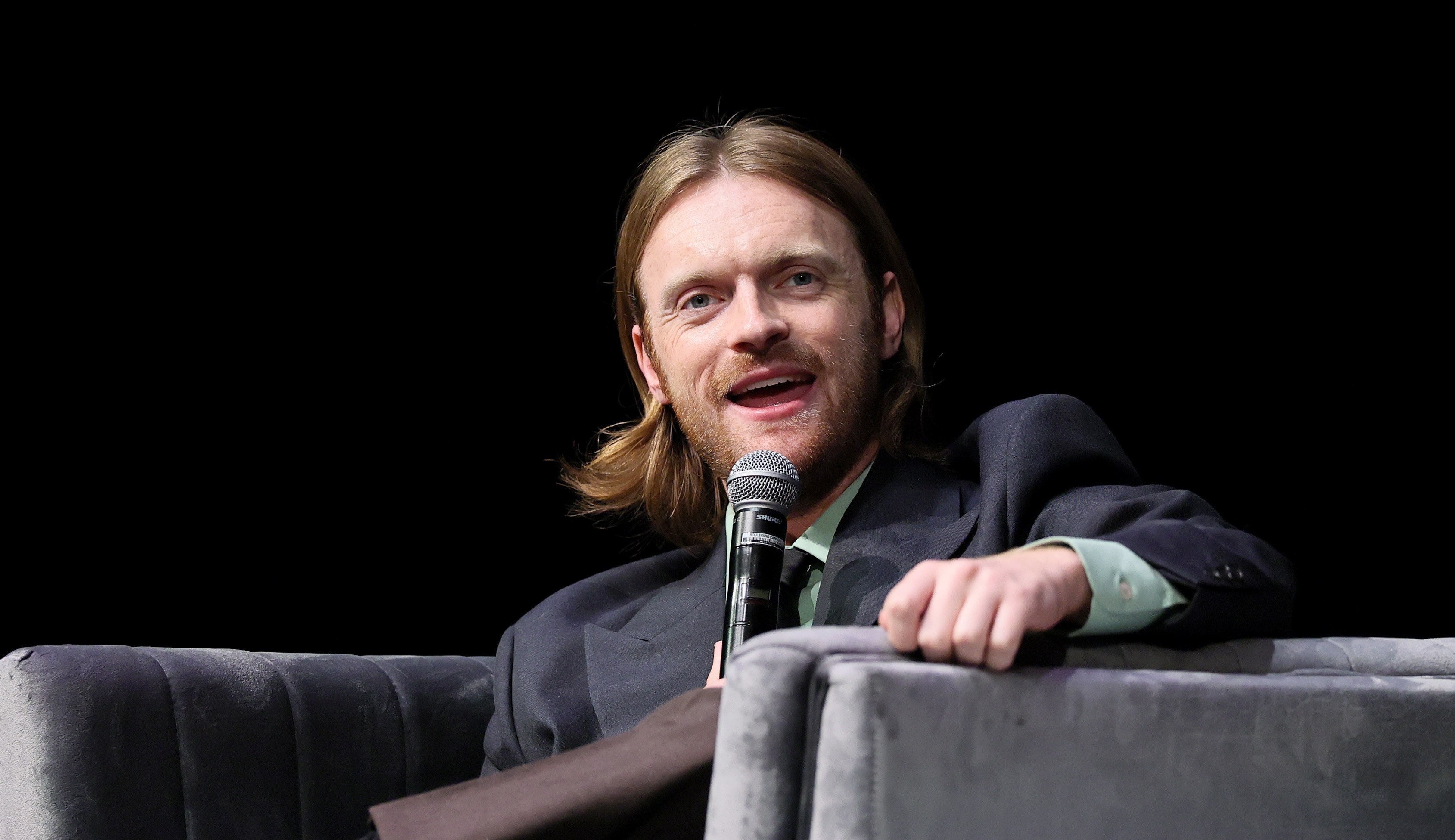 Finneas O'Connell speaking into a microphone while seated on a grey couch, wearing a dark jacket and light shirt.