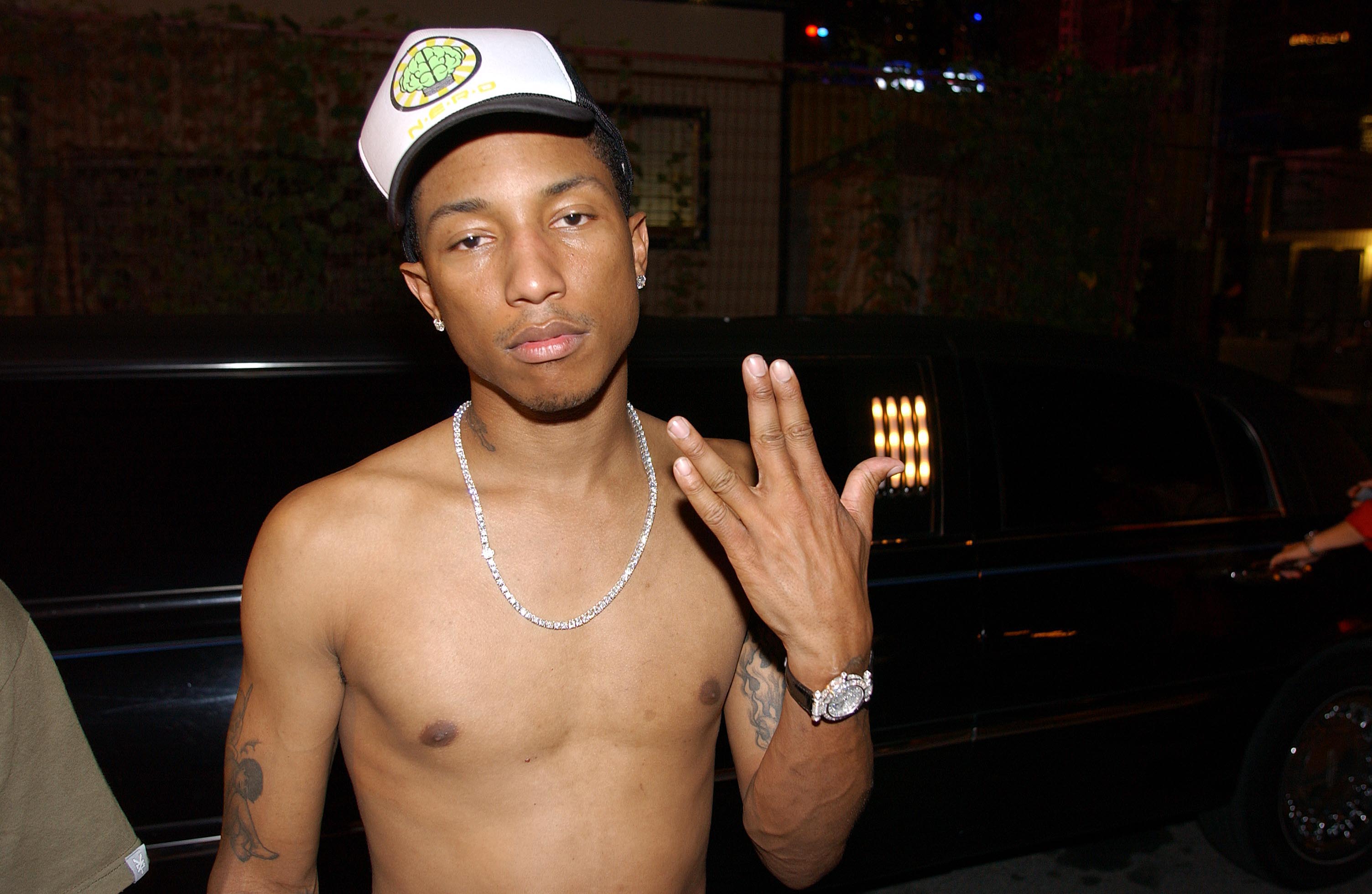 A shirtless Pharrell wearing a cap and chain necklace poses with a hand gesture in front of a black car.
