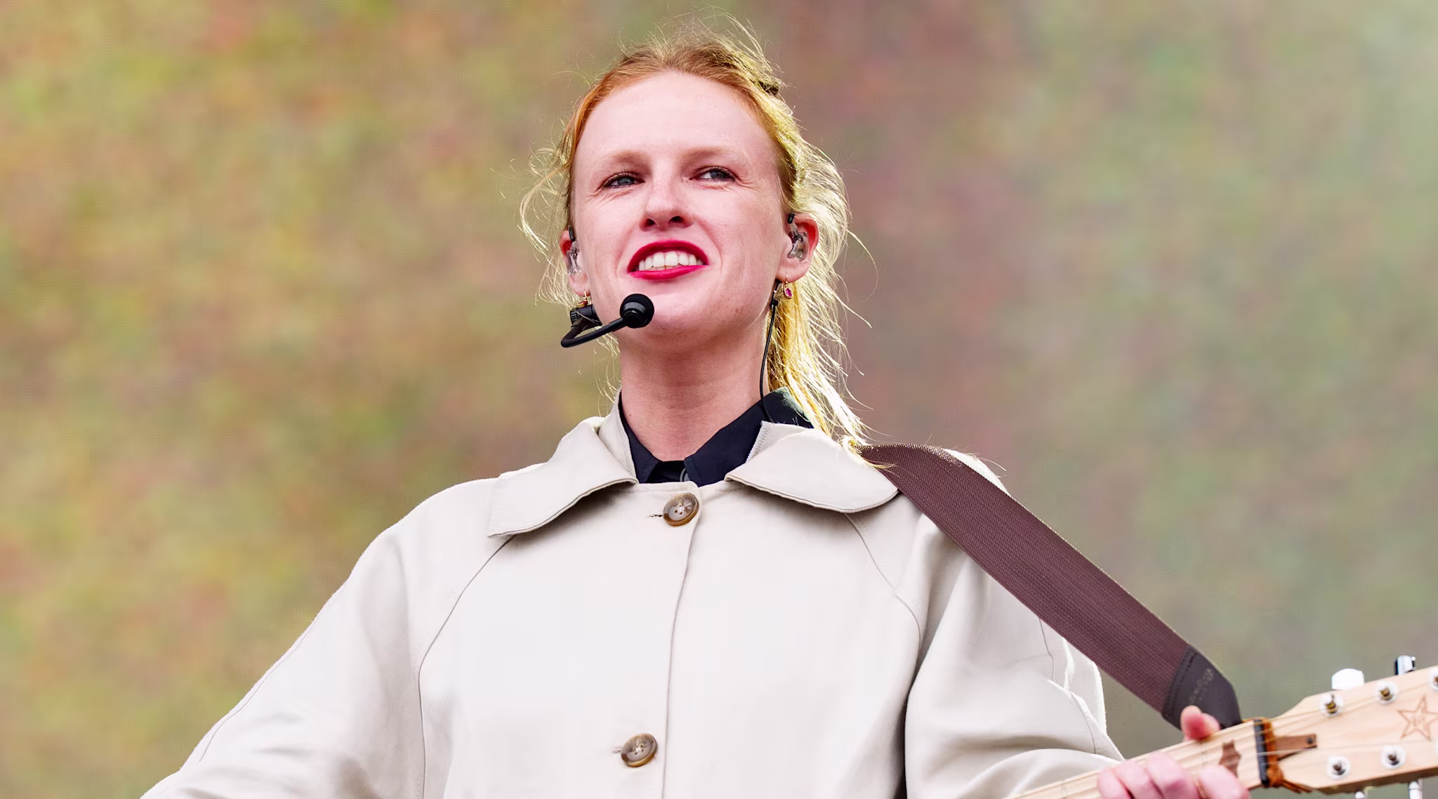 Audrey Hobert in a beige coat plays a banjo on stage, wearing a headset microphone. The background is a soft gradient.