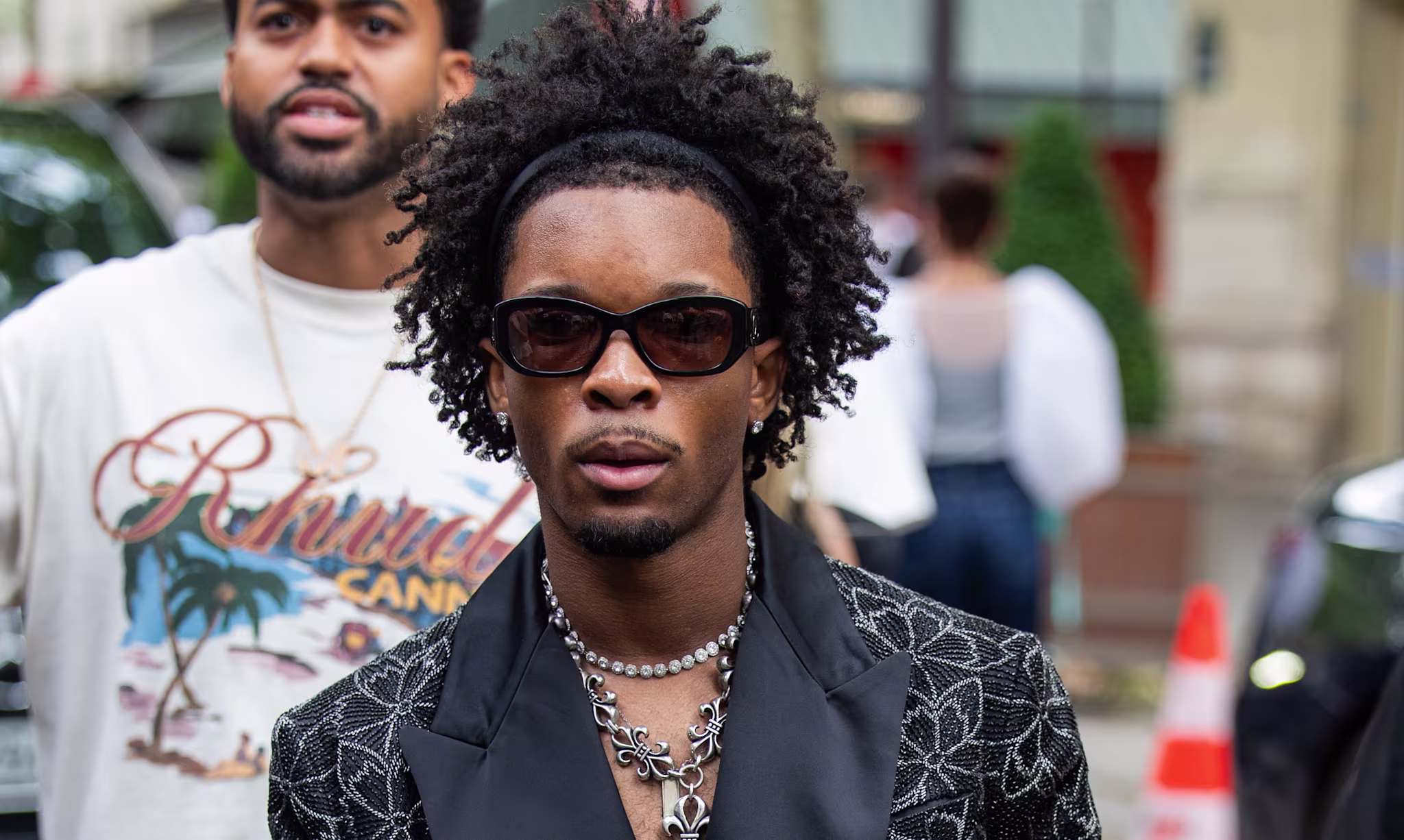 D4vd in sunglasses and a patterned black blazer with floral designs, wearing layered necklaces, walking on a city street.