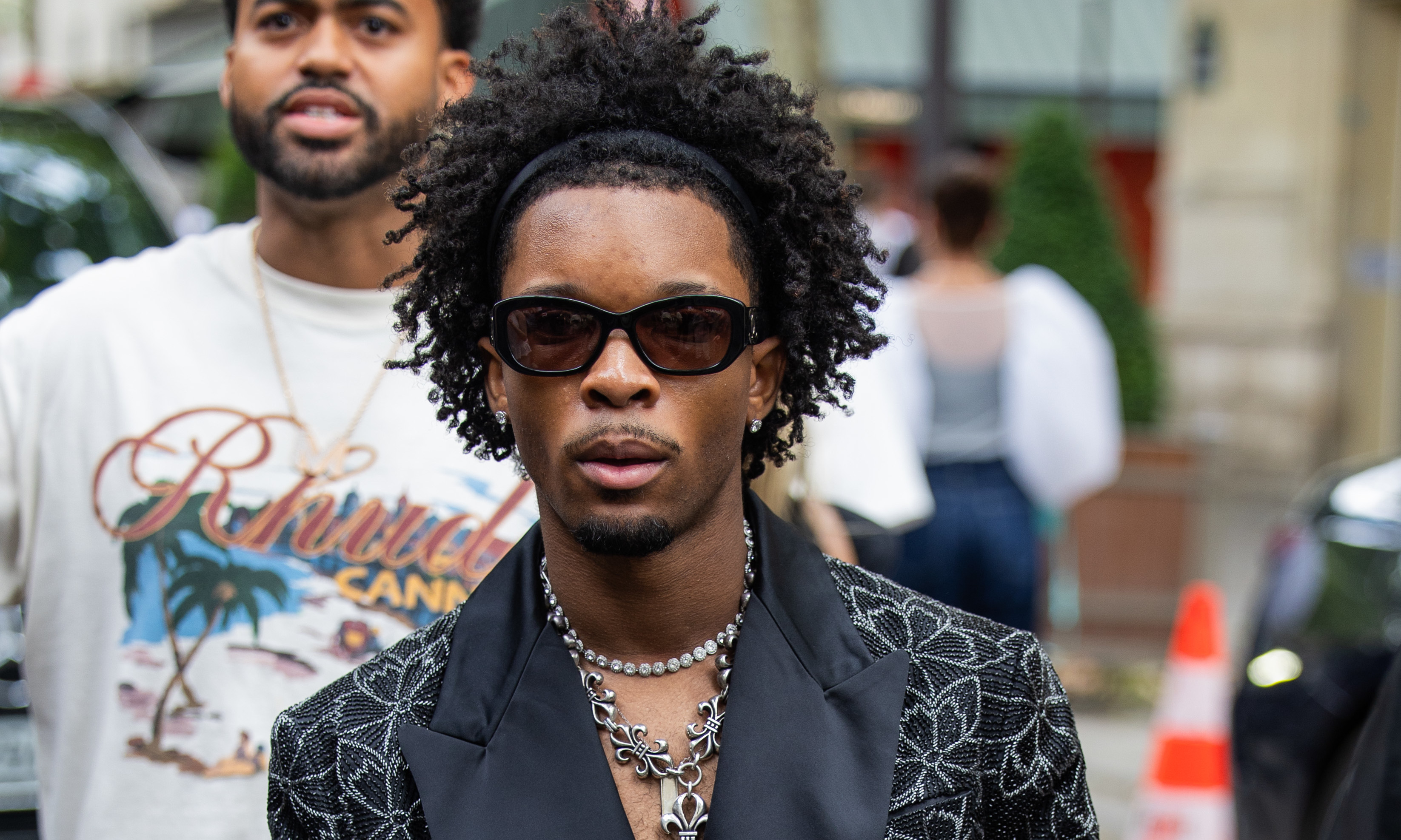 D4vd in sunglasses and a patterned black blazer with floral designs, wearing layered necklaces, walking on a city street.