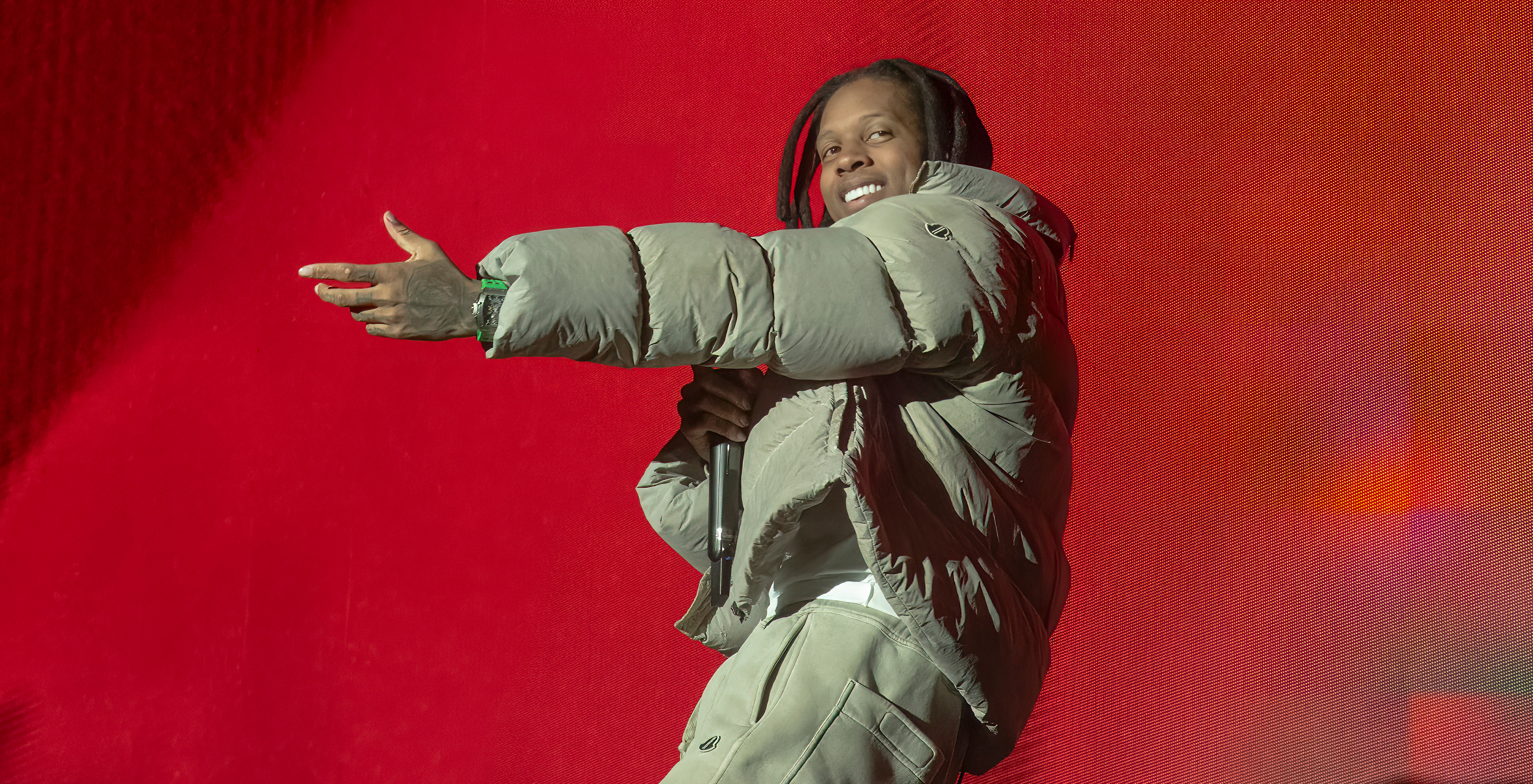 Lil Durk in a puffer jacket and pants holds a microphone on stage against a vibrant red background.