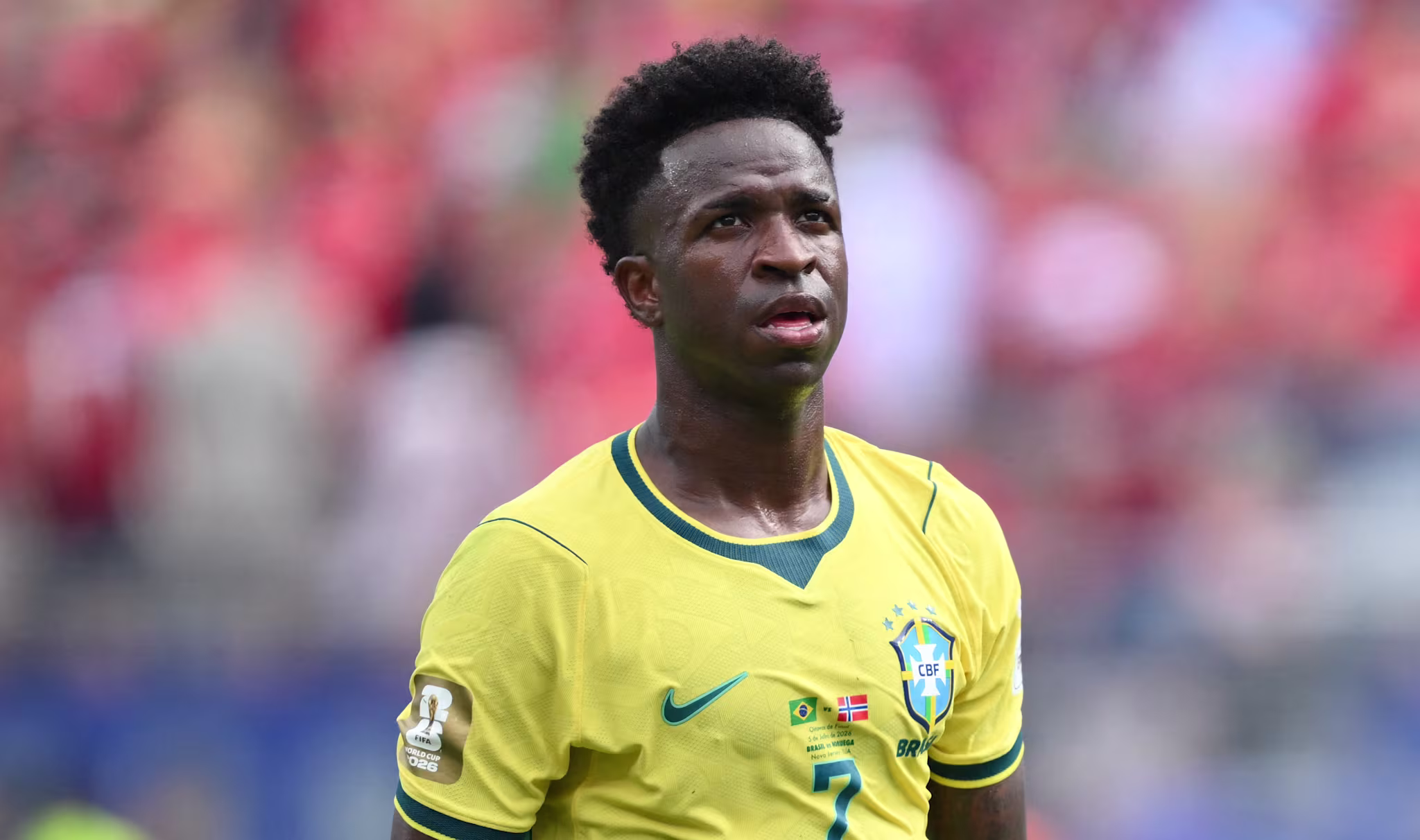 Vinícius Júnior in yellow jersey on the field, with a focused expression and blurred crowd in the background.