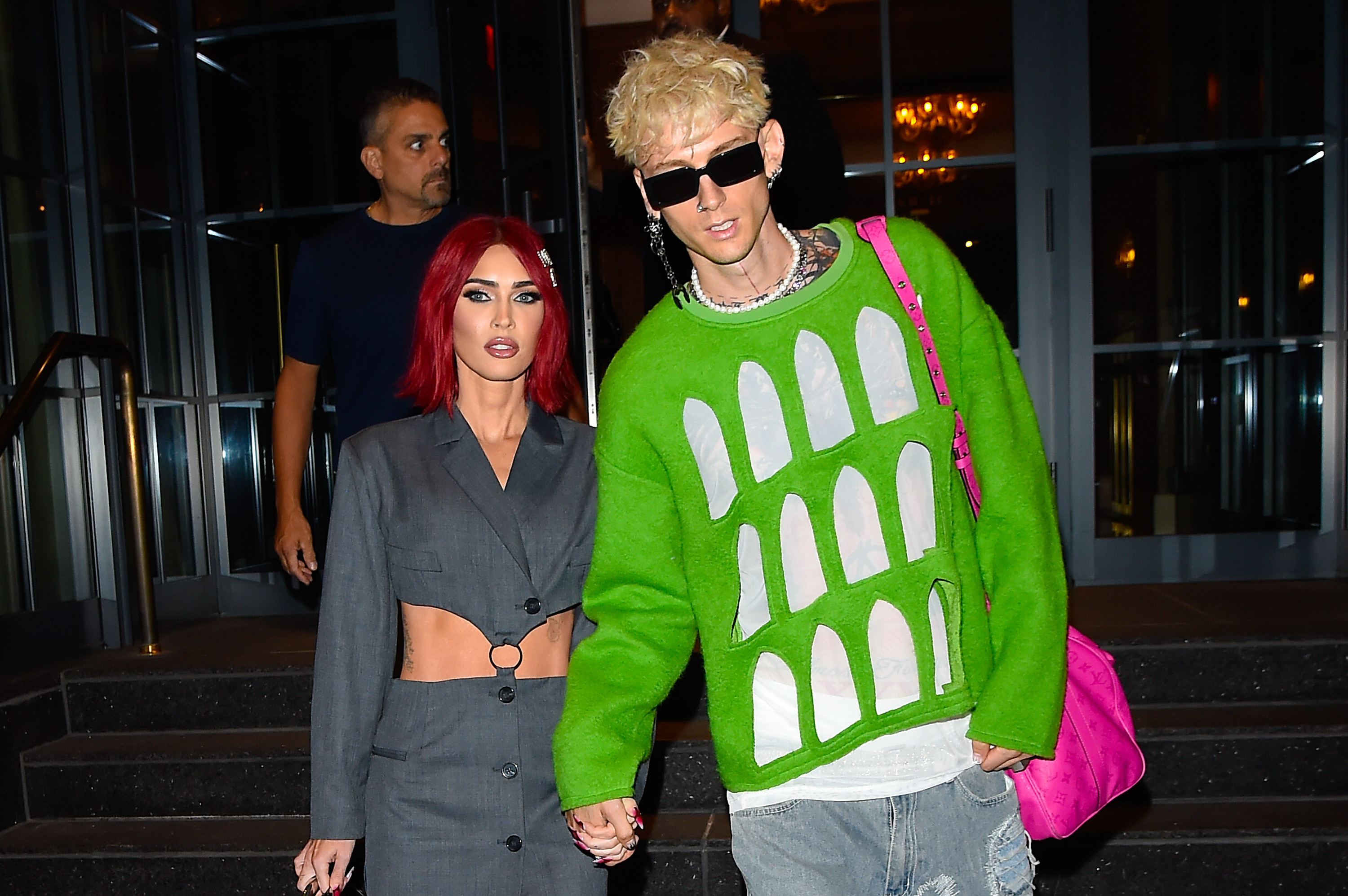 Megan Fox with red hair in a gray outfit and Machine Gun Kelly in a green sweater and sunglasses, holding hands outdoors.