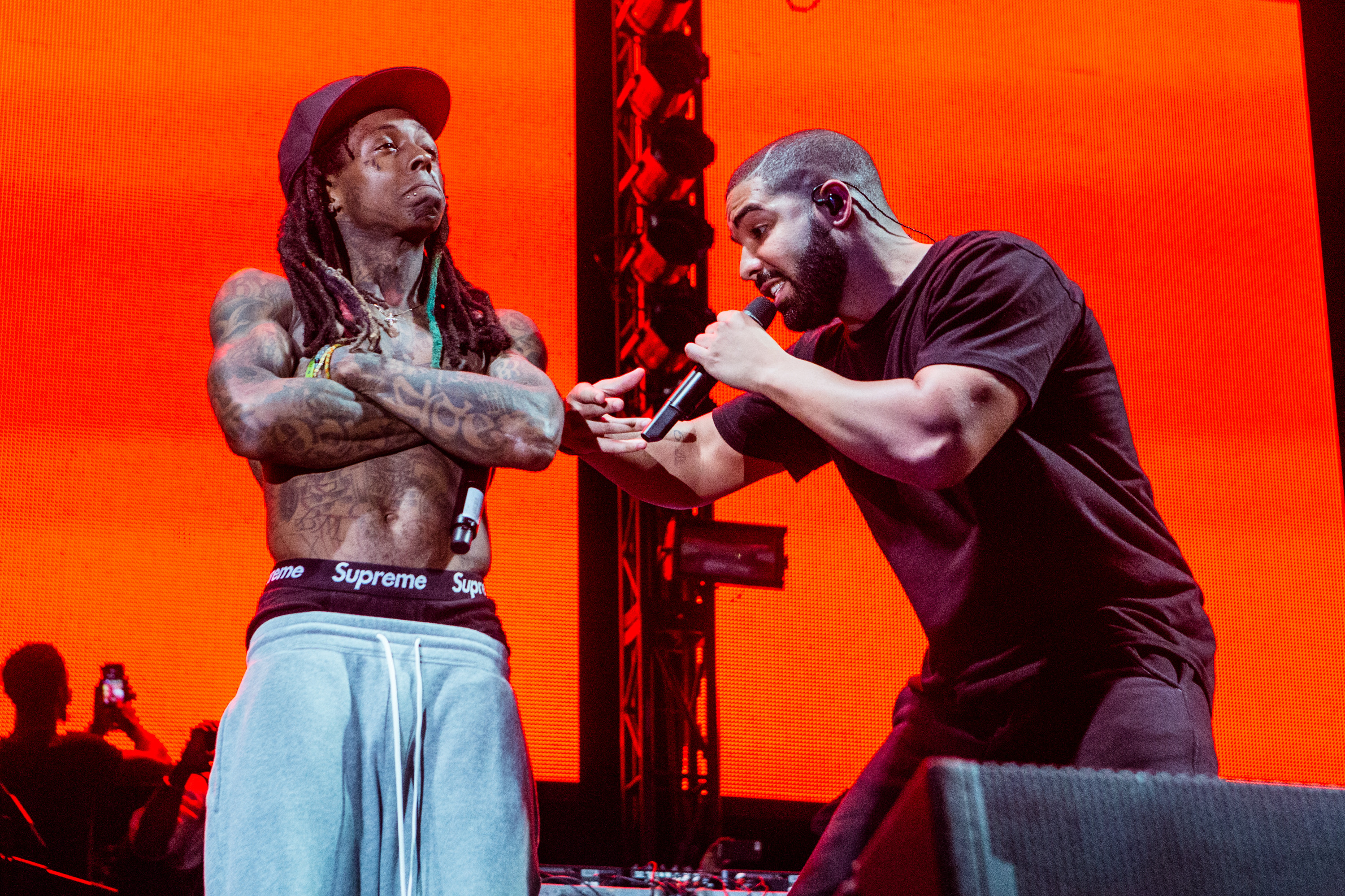 Lil Wayne and Drake performing in front of an orange backdrop. Lil Wayne has no shirt on and Drake is wearing a black shirt