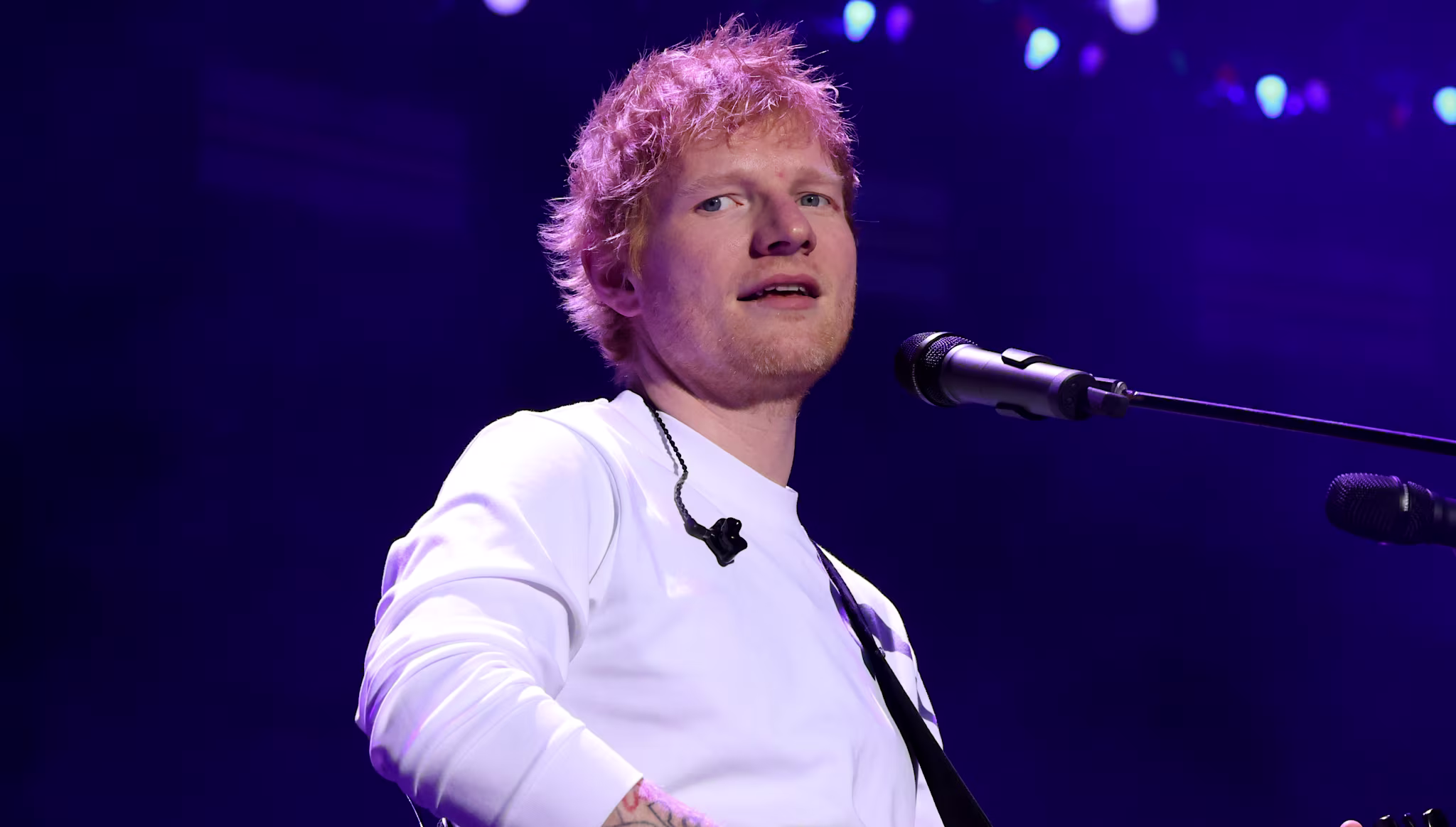Ed Sheeran performing on stage with a guitar, wearing a white shirt, under purple stage lights.