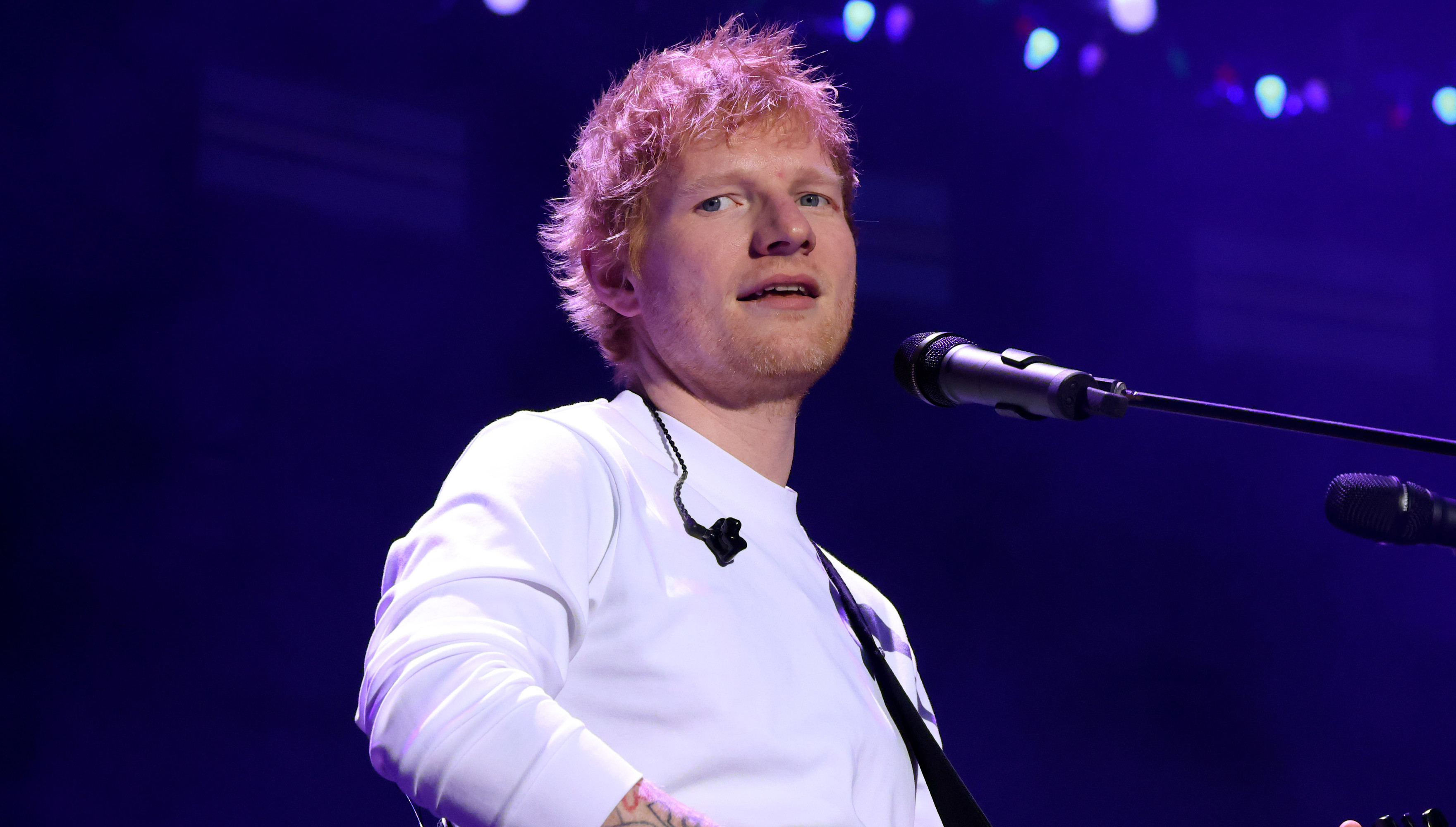 Ed Sheeran performing on stage with a guitar, wearing a white shirt, under purple lighting.