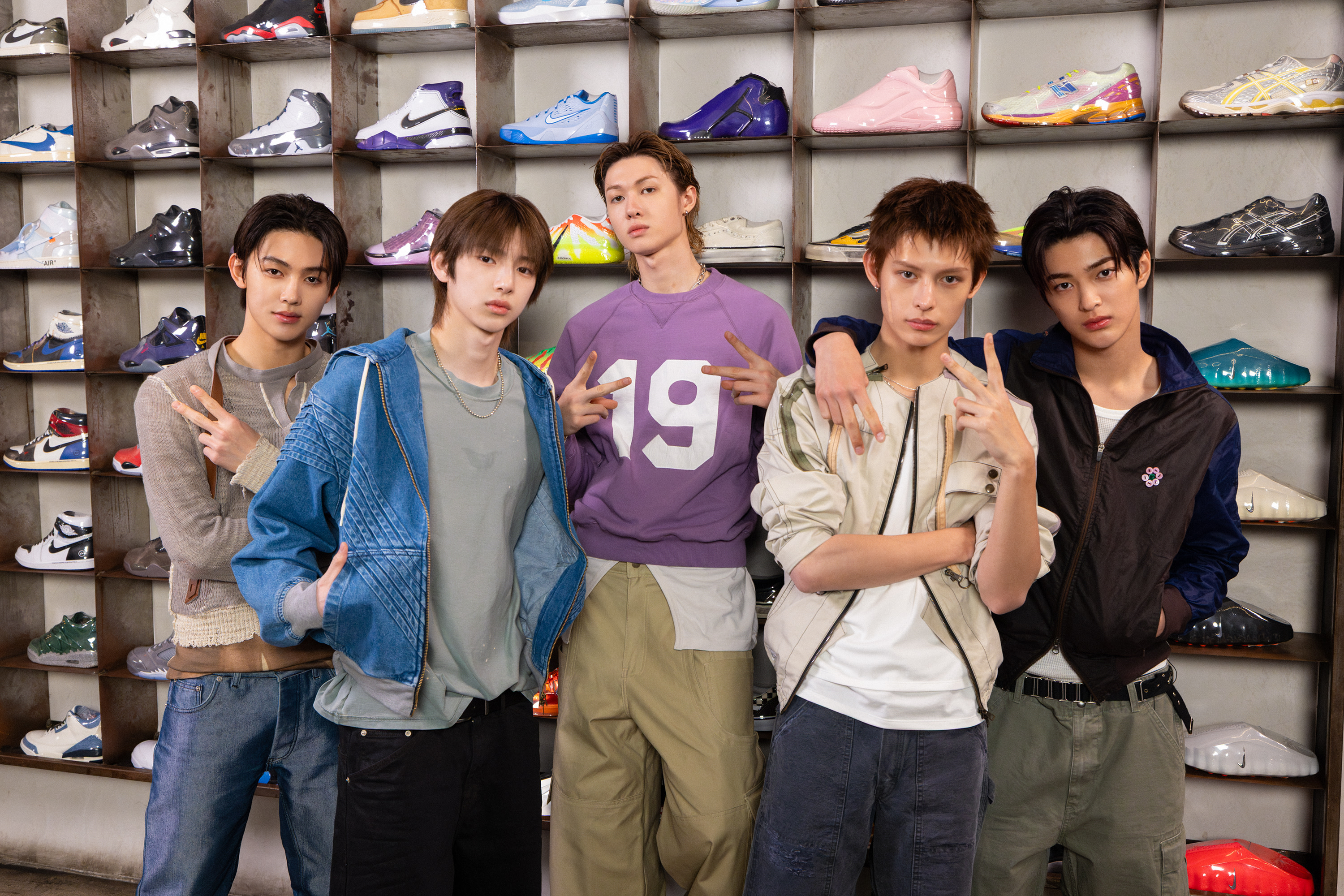 Five young men posing in front of a wall displaying various sneakers, making peace signs and casual expressions.