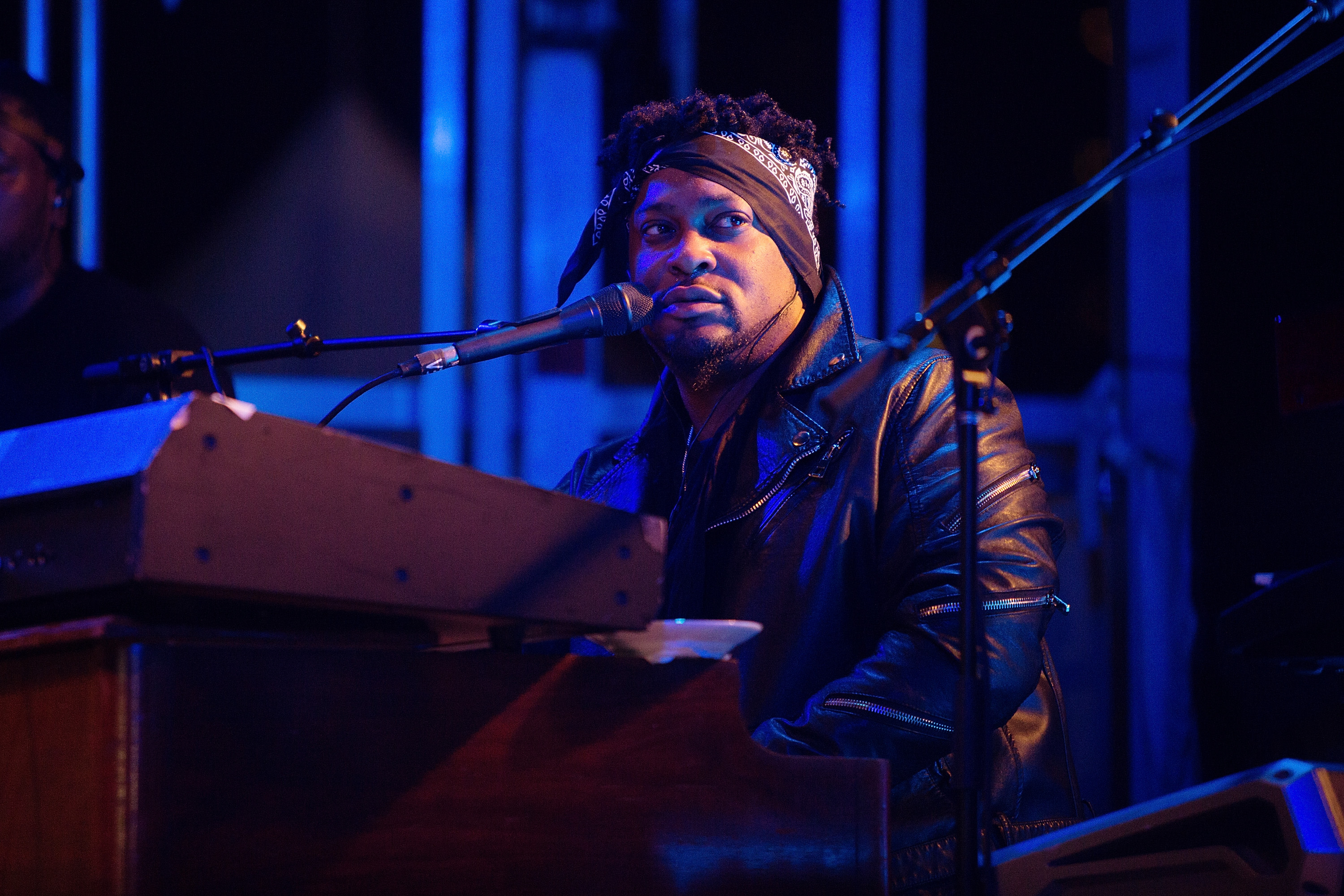 A musician wearing a leather jacket and headband plays the keyboard on stage, illuminated by blue lighting.