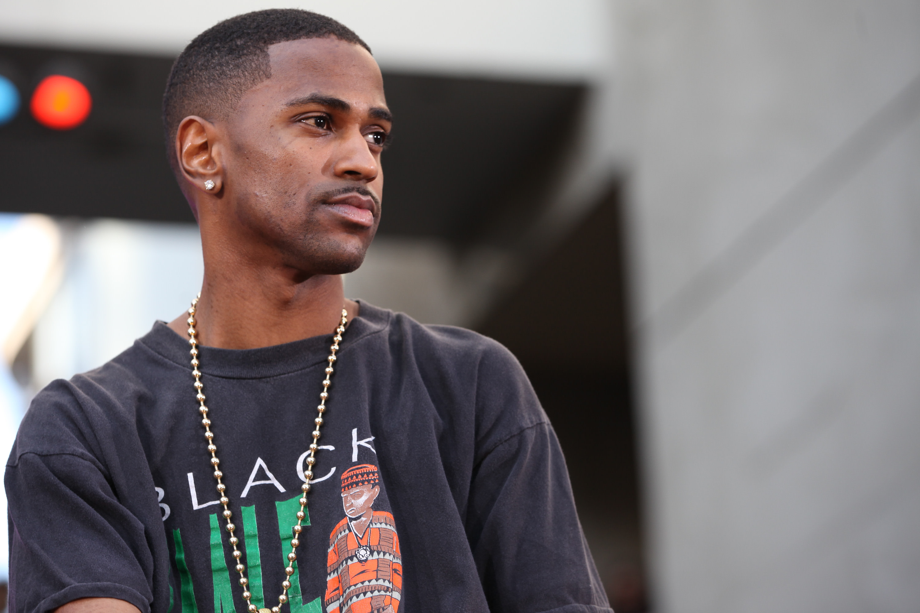 Big Sean wearing a black graphic T-shirt and a gold chain, standing outdoors with a neutral expression.