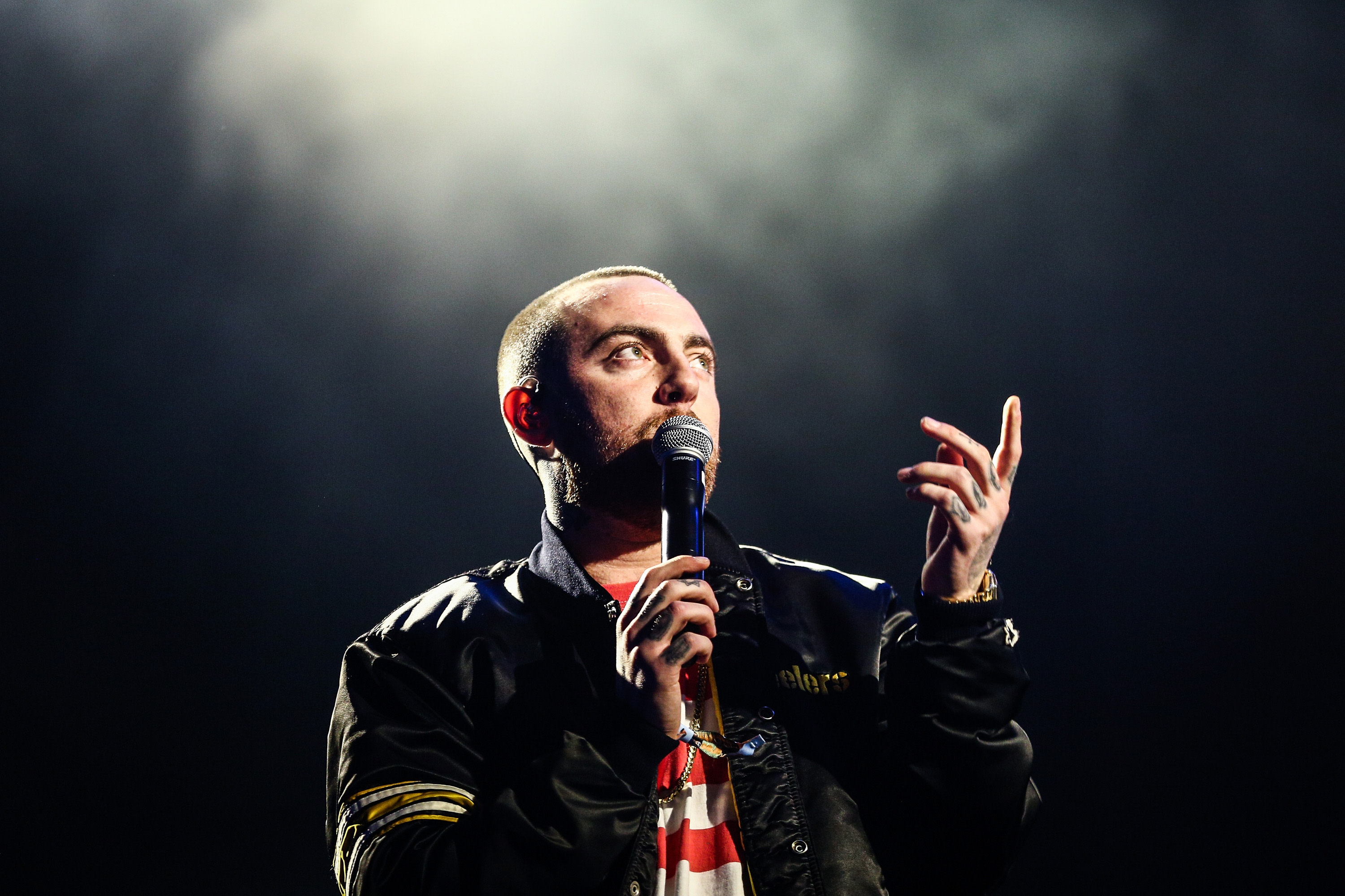 Rapper Mac Miller performing on stage, holding a microphone, wearing a dark jacket with a focused expression.