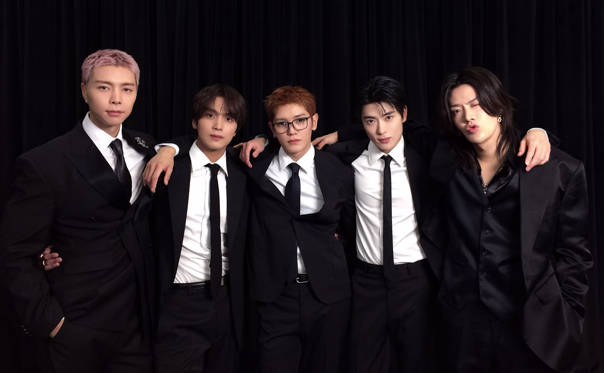Five men in black suits and ties pose together against a black curtain backdrop.