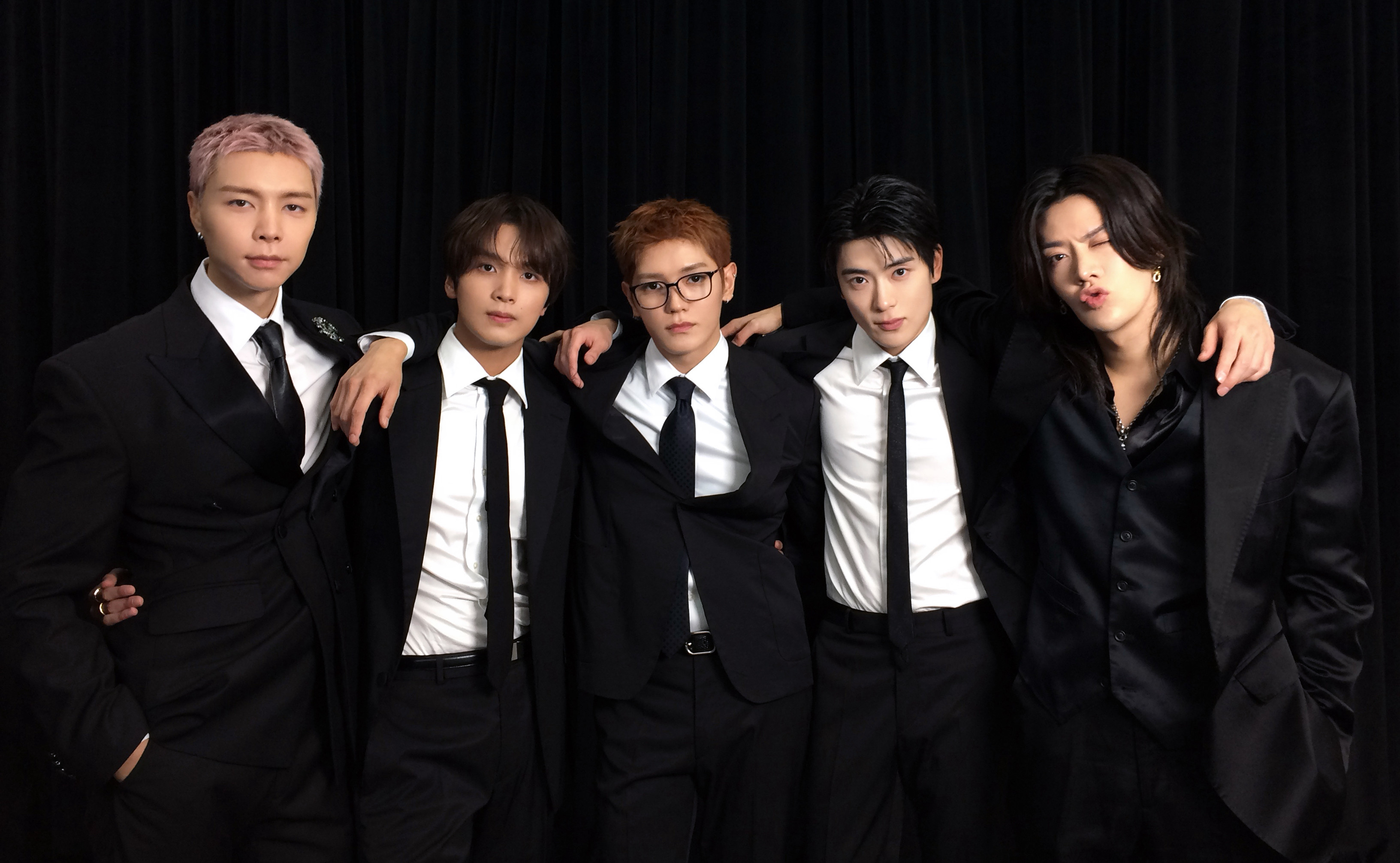 Five men in black suits and ties pose together against a black curtain backdrop.