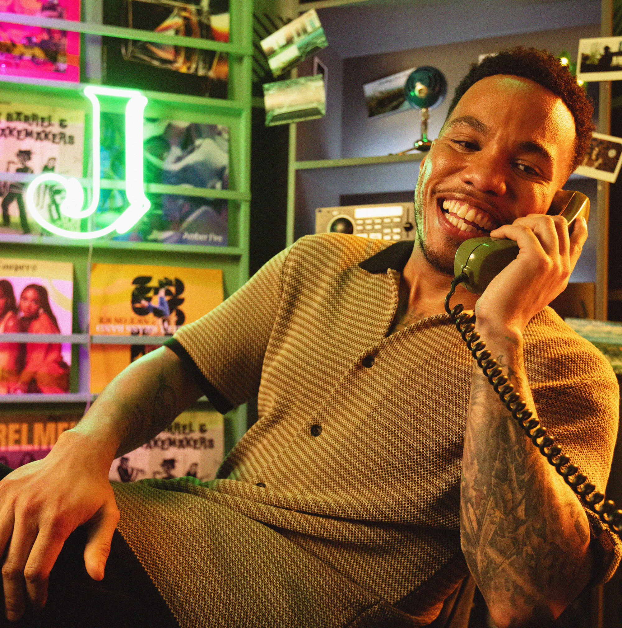 A man smiling and talking on a vintage phone, sitting in a room with colorful records and a neon "J" sign in the background.