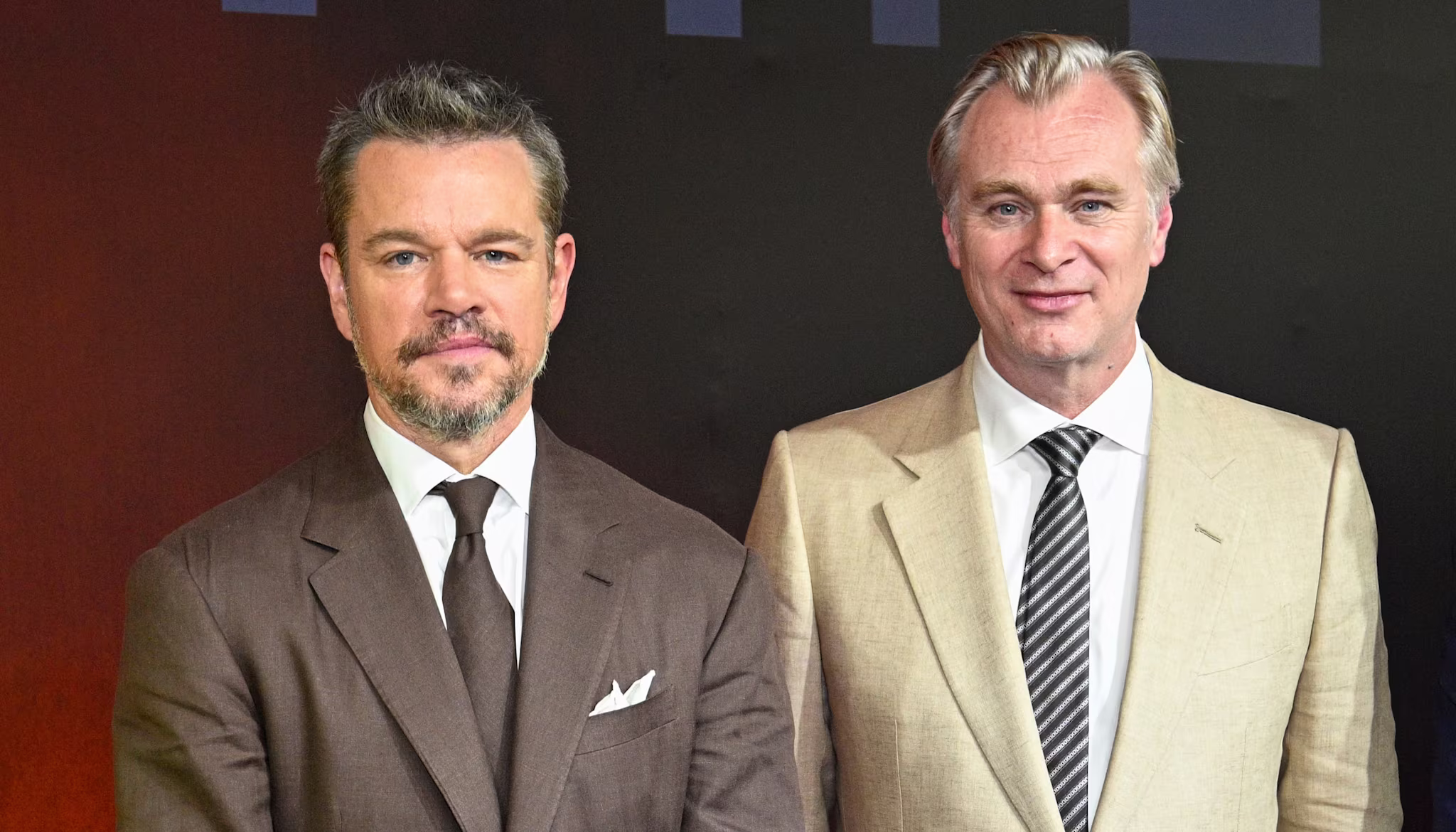 Matt Damon and Christopher Nolan on a red carpet. Damon in a brown suit, Nolan in beige.