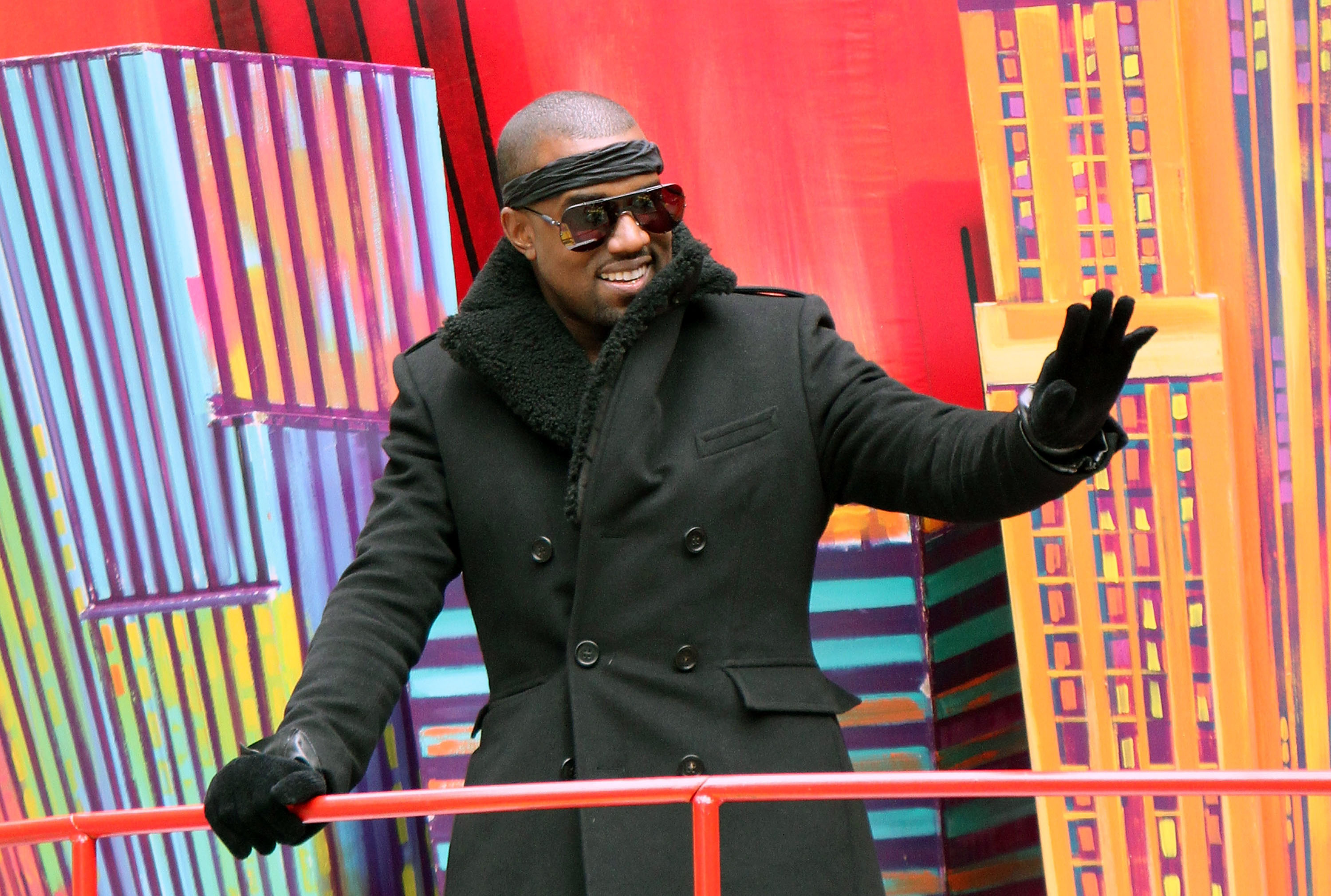 Kanye West in a black coat and sunglasses, waving, with a colorful cityscape background.