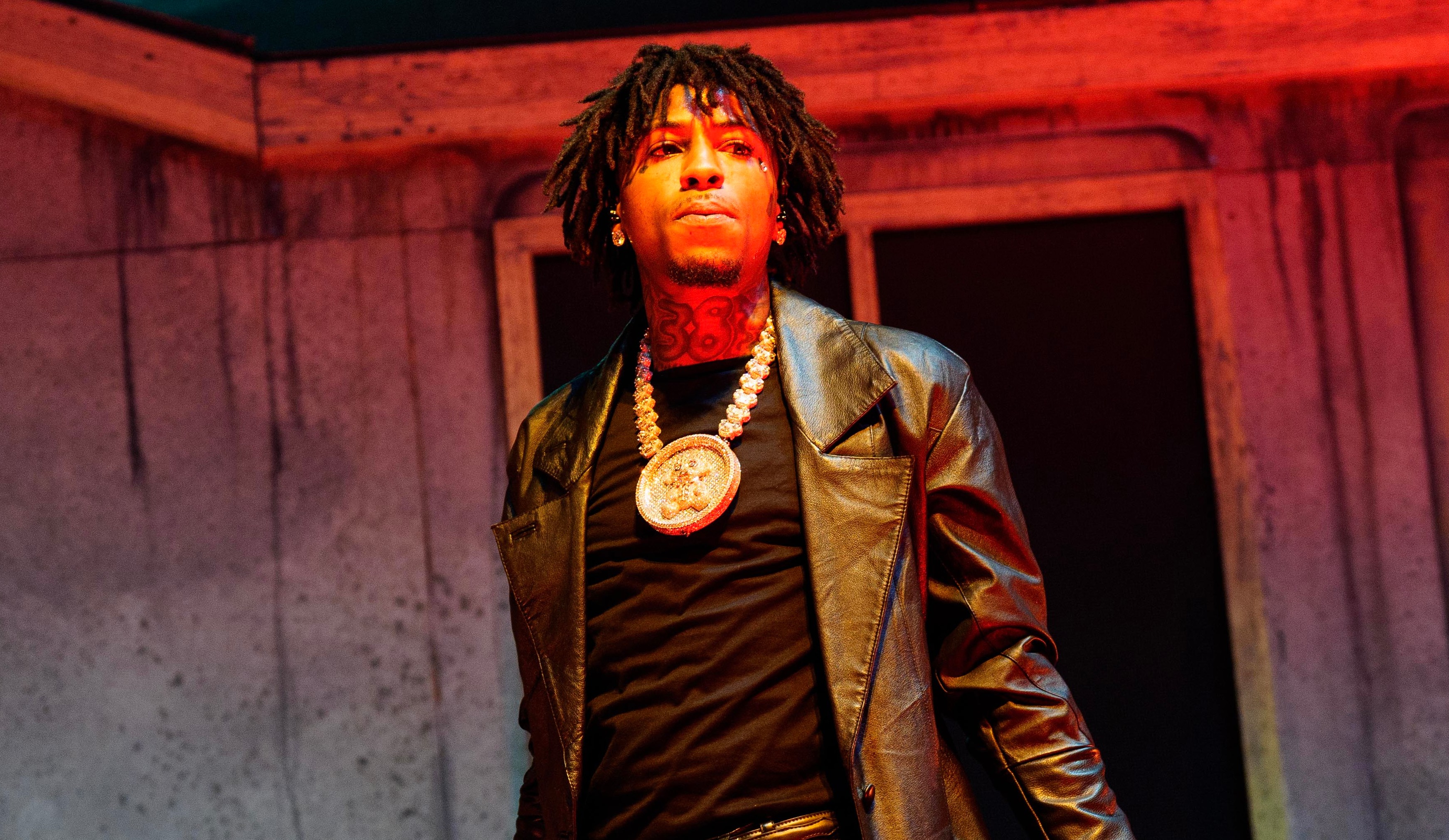 NBA Youngboy in a black outfit and long coat stands in front of a building, wearing a large pendant and holding a microphone.