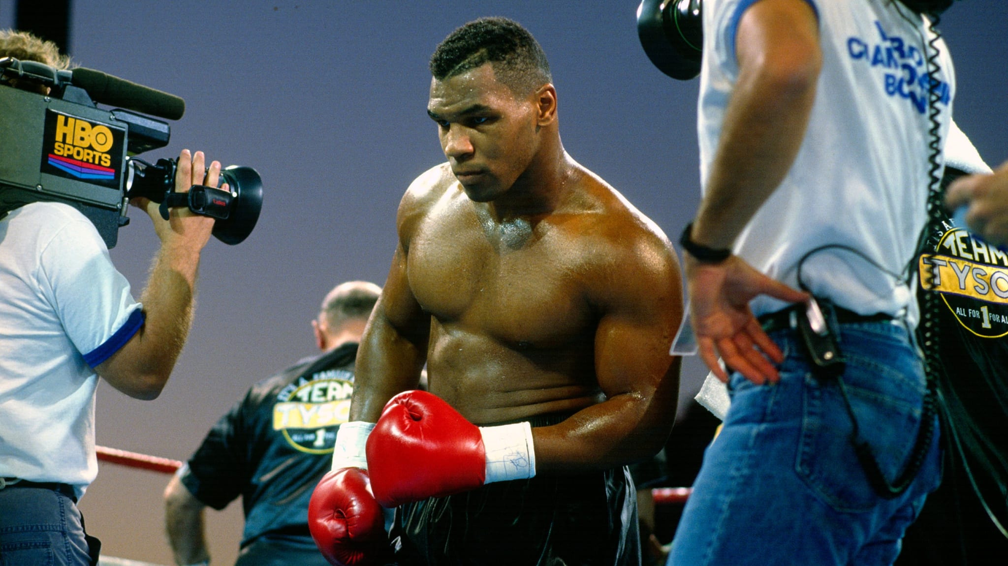 Mike Tyson, wearing red boxing gloves, is in a boxing ring surrounded by cameras and crew.