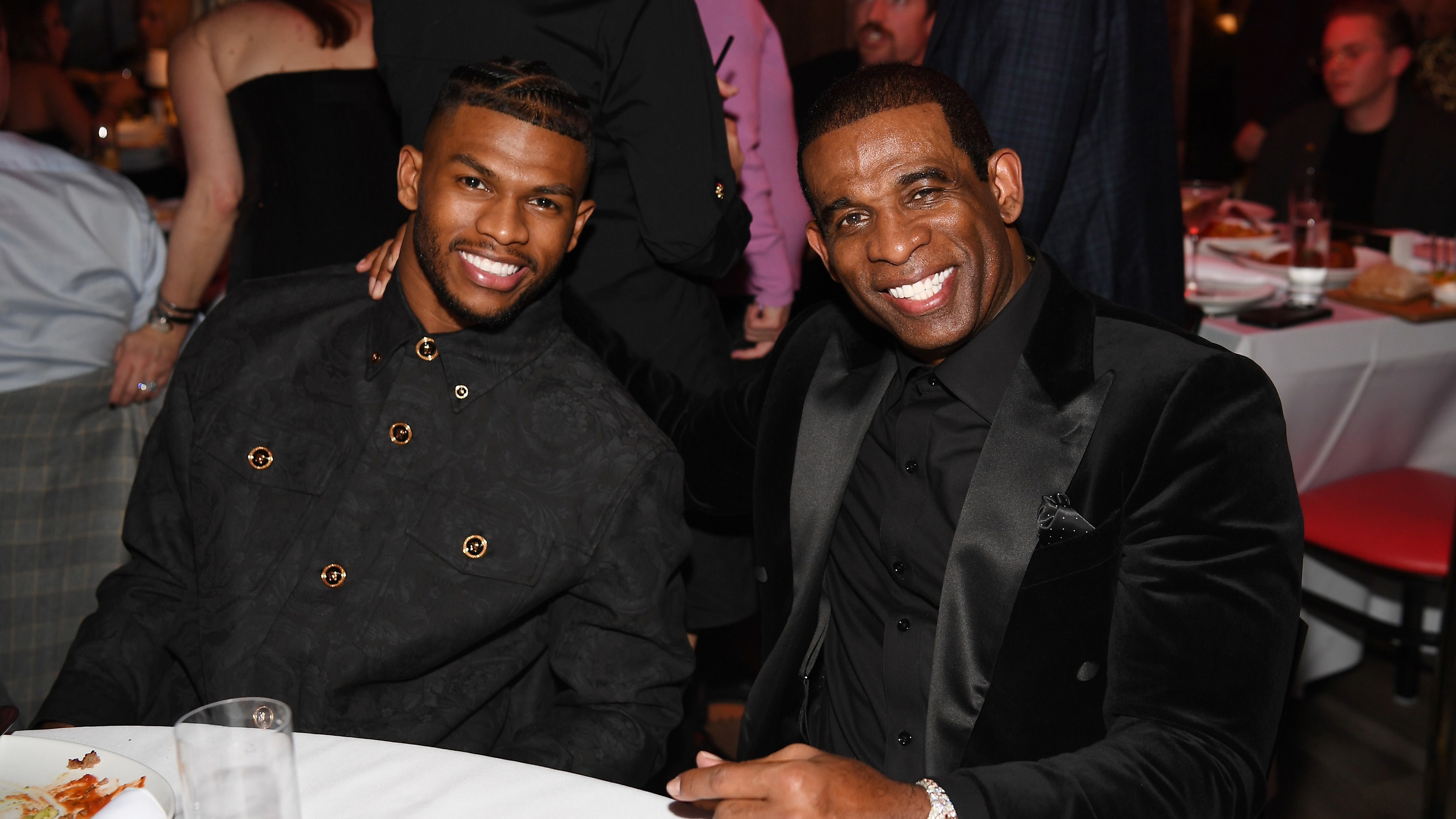 Deion Sanders and Shiloh Sanders at a formal event, seated at a table. One is wearing a black jacket, the other a black suit.
