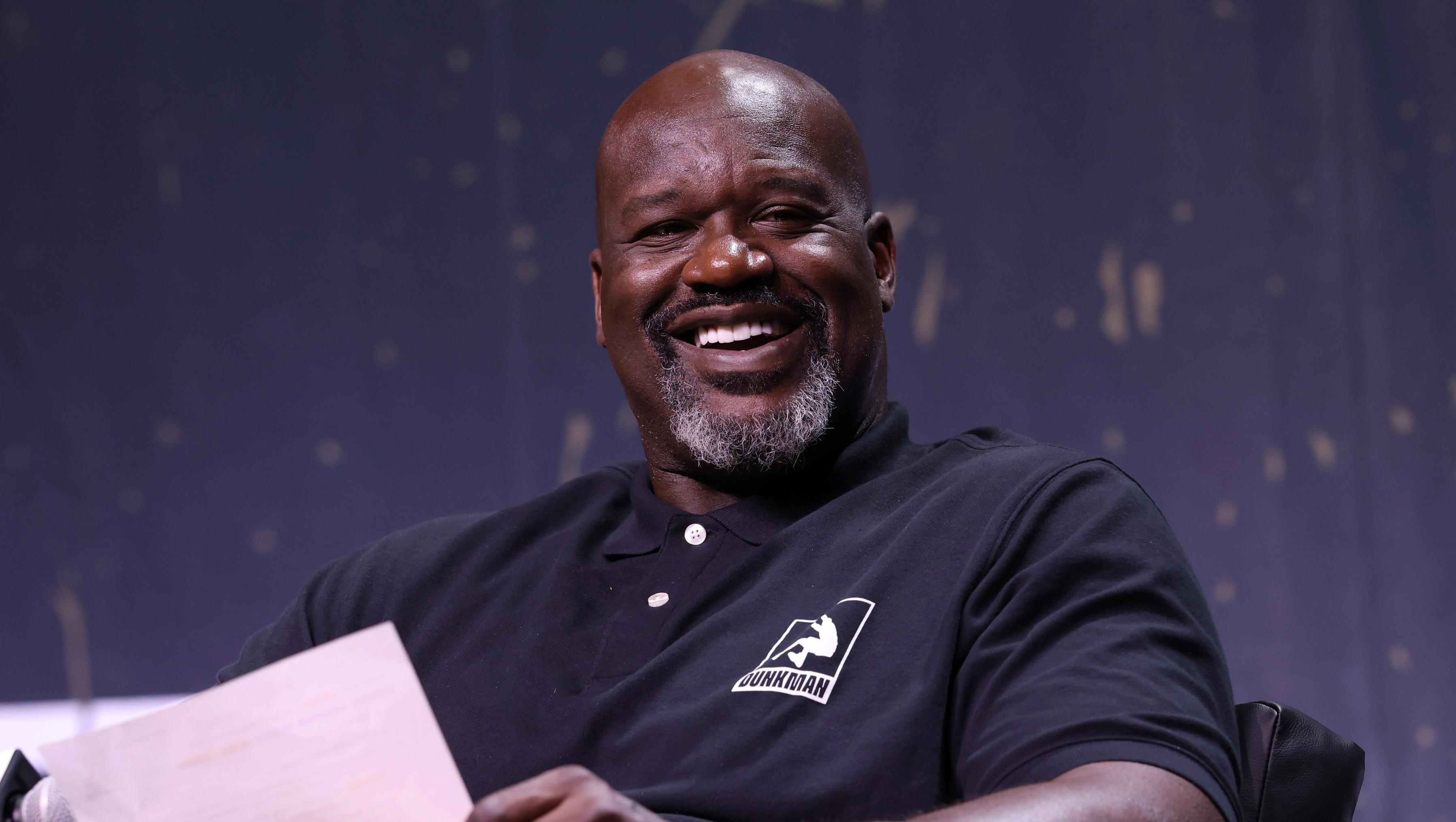 Shaquille O'Neal smiling, seated on a stage, wearing a black polo shirt and khaki pants, holding a paper.