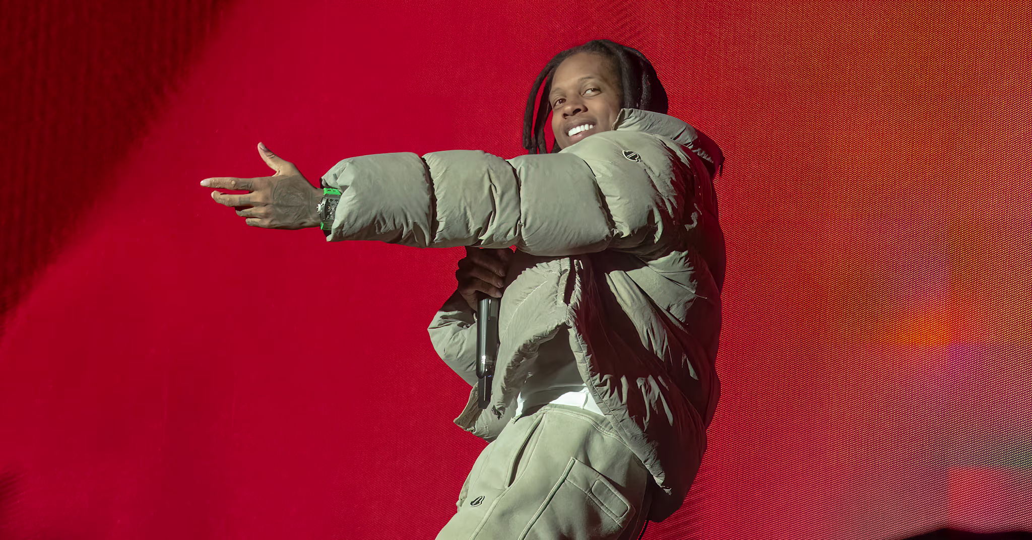 Lil Durk in a puffer jacket and pants holds a microphone on stage against a vibrant red background.