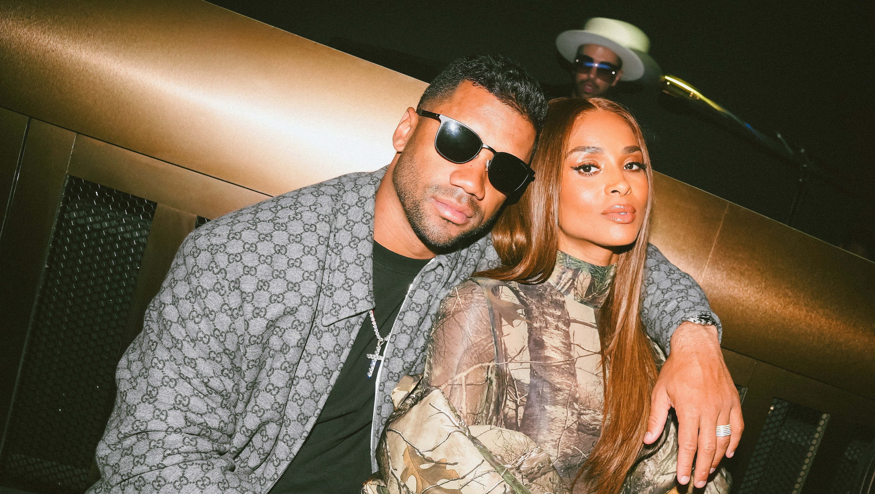 Russell Wilson in sunglasses and a patterned jacket sits with Ciara in a camouflage outfit holding a drink, in a dimly lit setting.