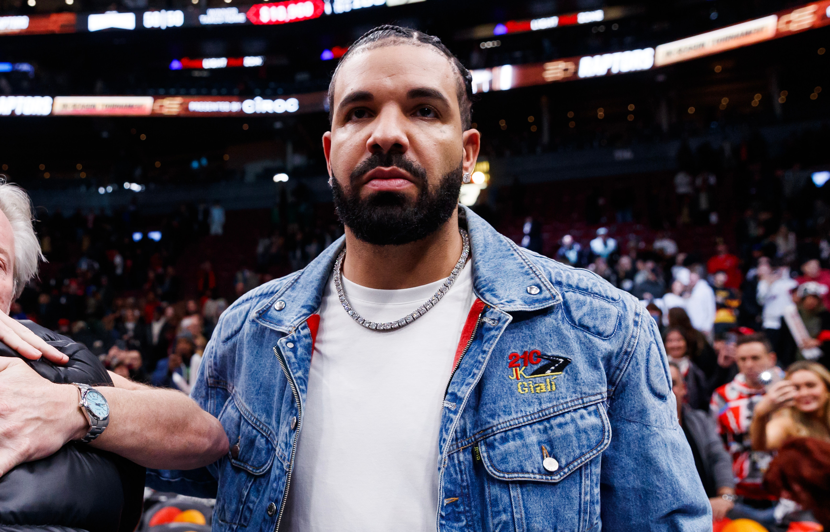 Drake in a denim jacket stands courtside at a basketball game, while two people hug nearby. Stadium seats are visible in the background.