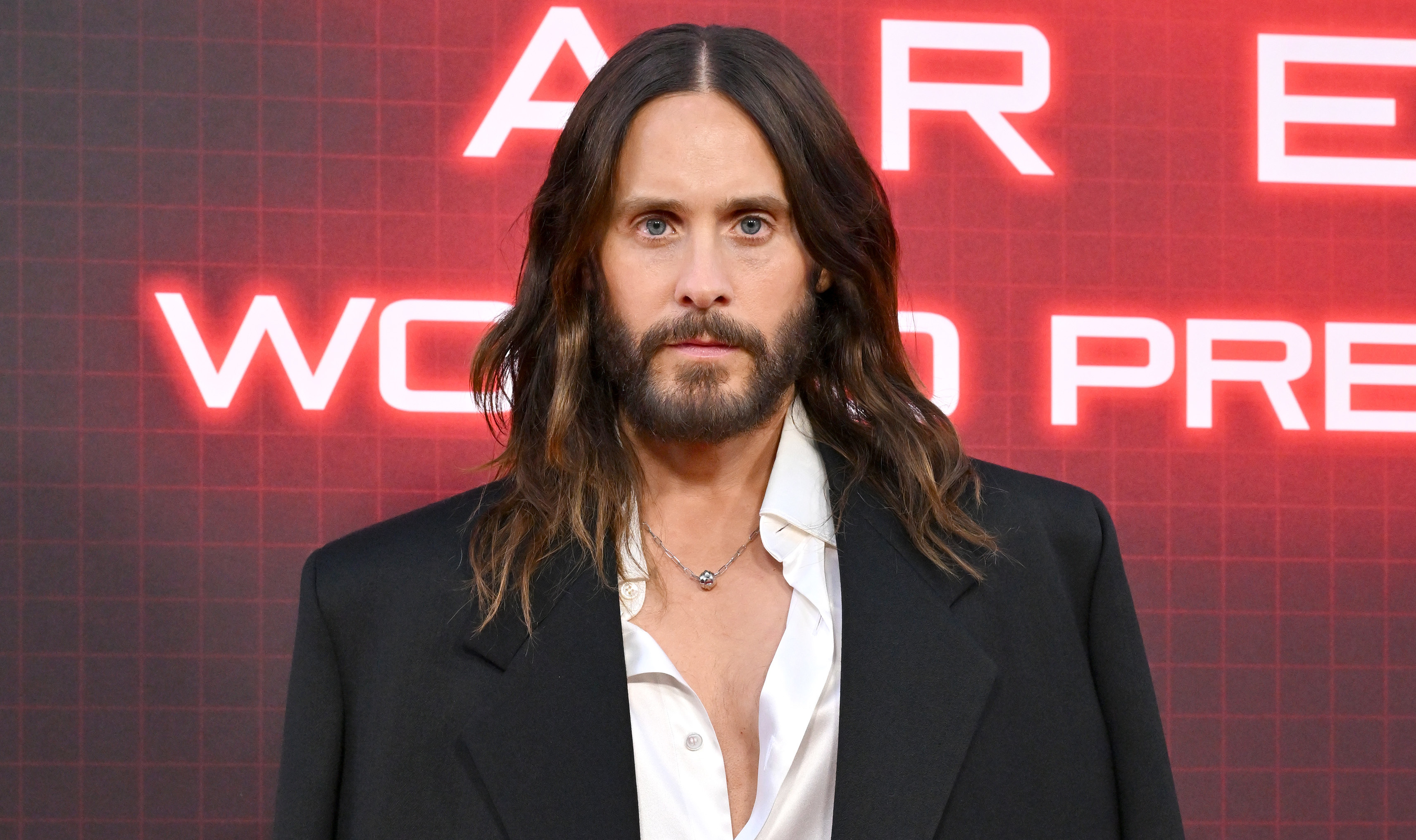 Jared Leto at a premiere, wearing a black oversized blazer over a white shirt, with long hair and a beard, standing against a red backdrop.