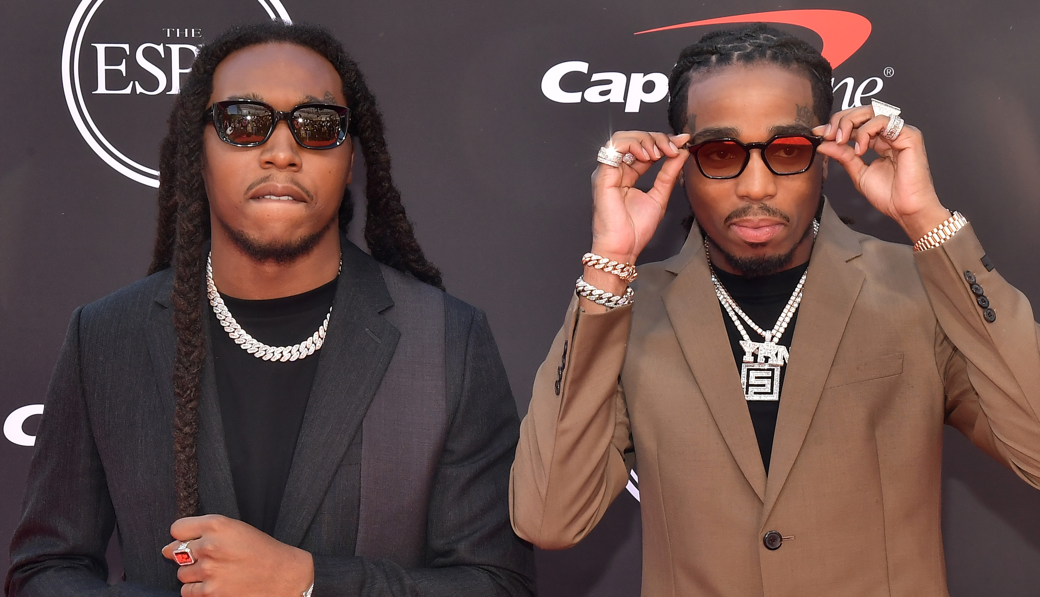 Takeoff and Quavo at the ESPY Awards, wearing suits and sunglasses, with jewelry and a backdrop featuring sponsor logos.