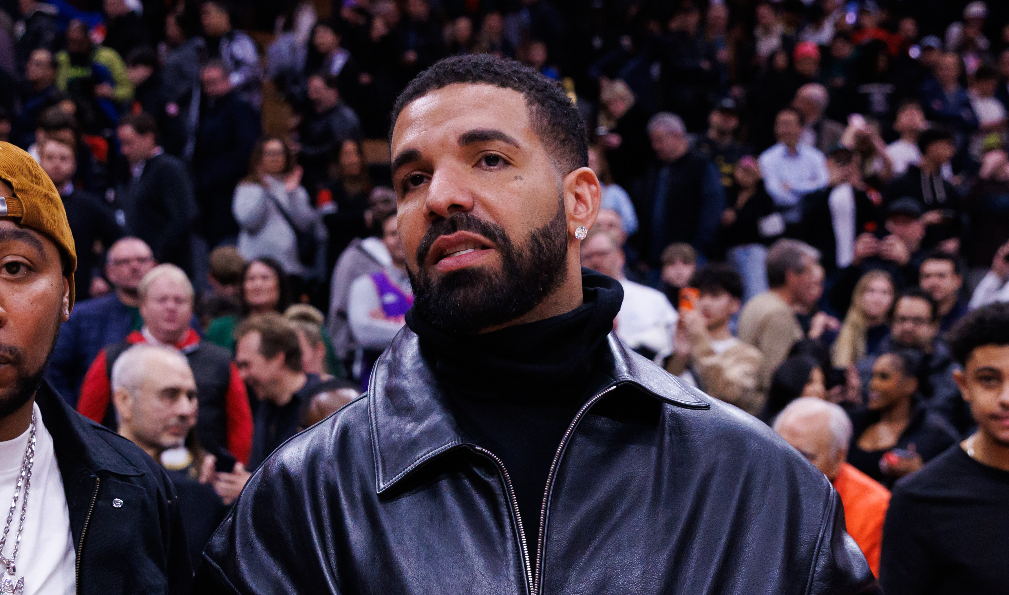 Drake in a black leather jacket at a sports event, surrounded by a crowd.