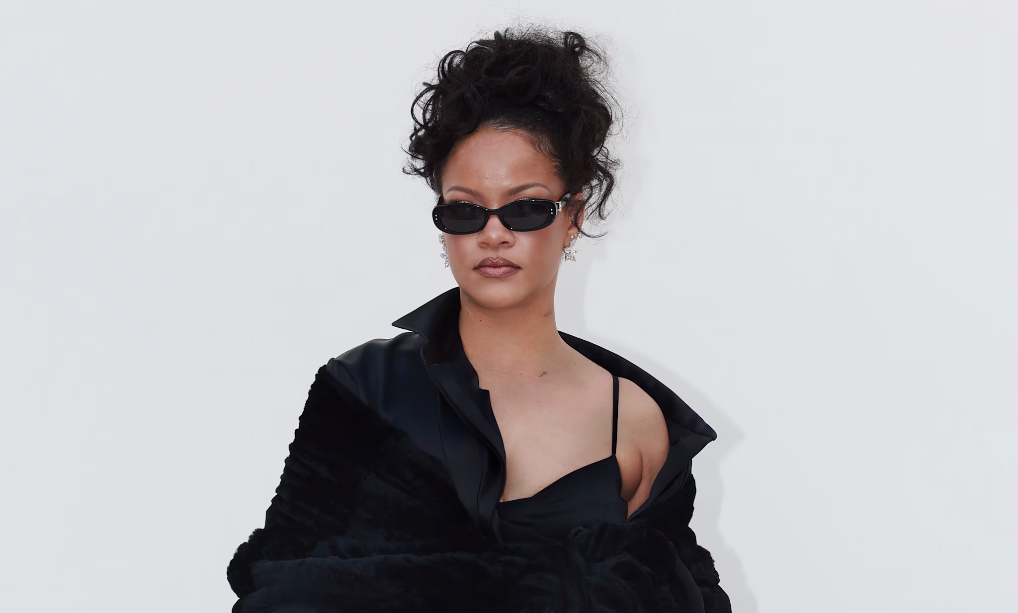 Rihanna in a stylish black outfit with sunglasses, showcasing tattoos and wearing high heels.