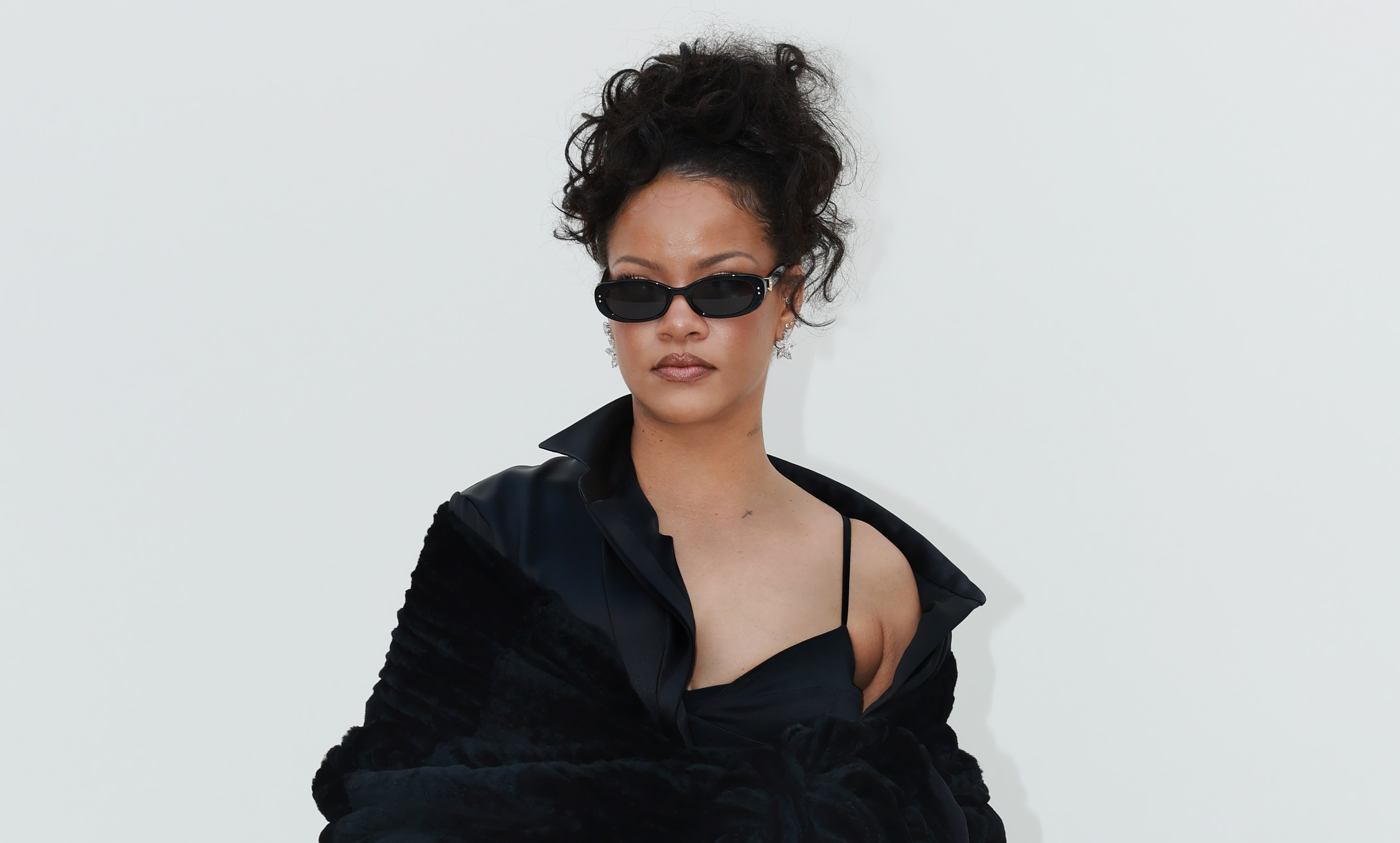 Rihanna in a stylish black outfit with sunglasses, showcasing tattoos and wearing high heels.