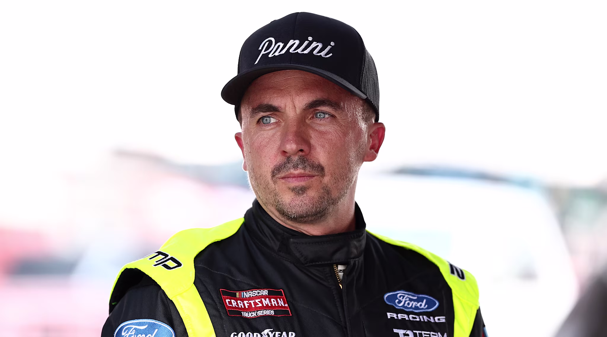 Frankie Muniz in a racing suit and cap with "Panini" logo, standing outdoors. The suit has various sponsor logos.