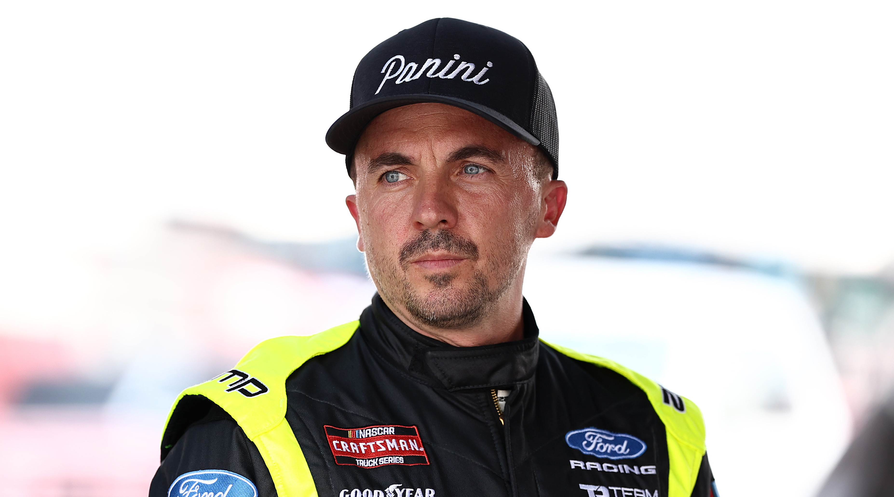 Frankie Muniz in a racing suit and cap with "Panini" logo, standing outdoors. The suit has various sponsor logos.