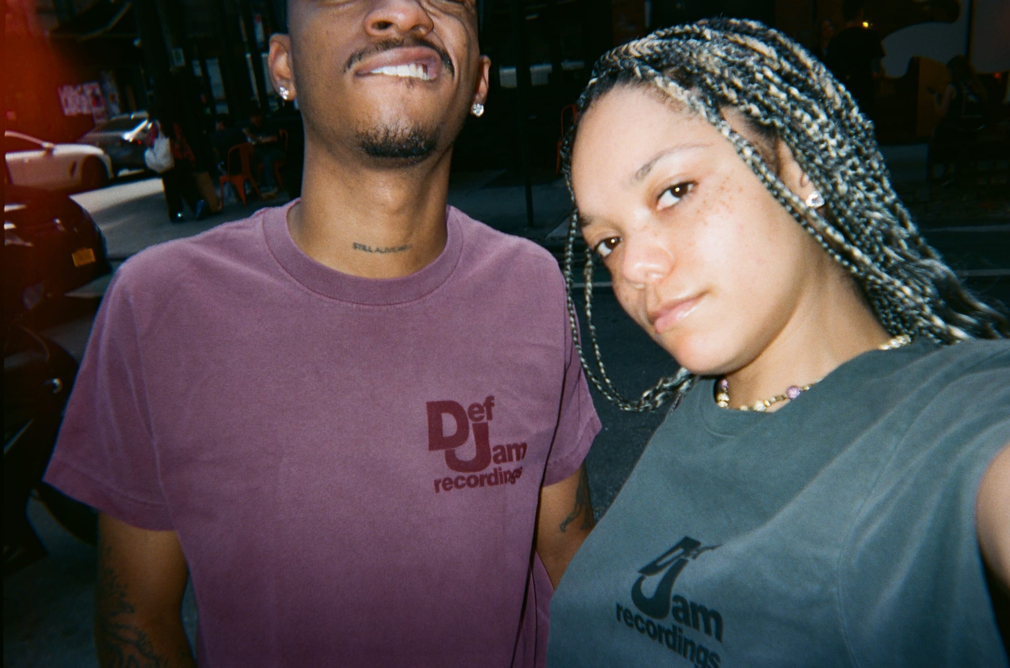 Two people taking a selfie, both wearing Def Jam Recordings shirts. The person on the right has braided hair.