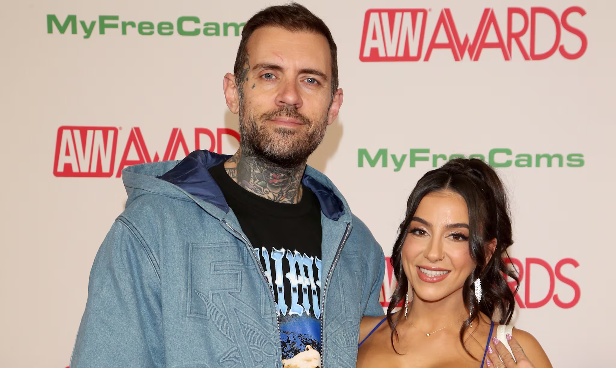 Adam22 in a blue jacket and jeans stands next to Lena The Plug in a sparkling blue dress at the AVN Awards event.