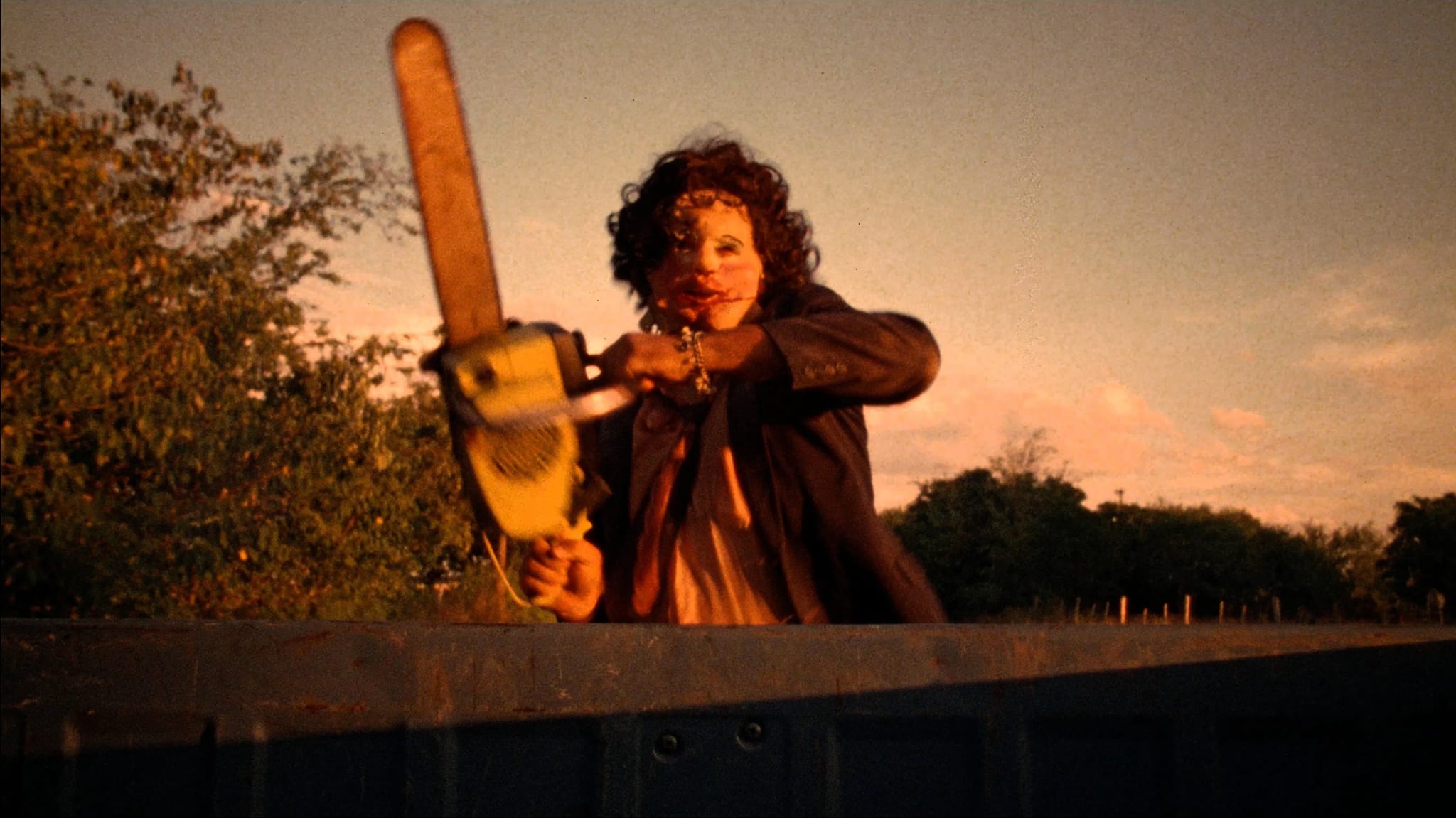 A person in a mask wielding a chainsaw stands behind a barrier at sunset, with trees in the background.