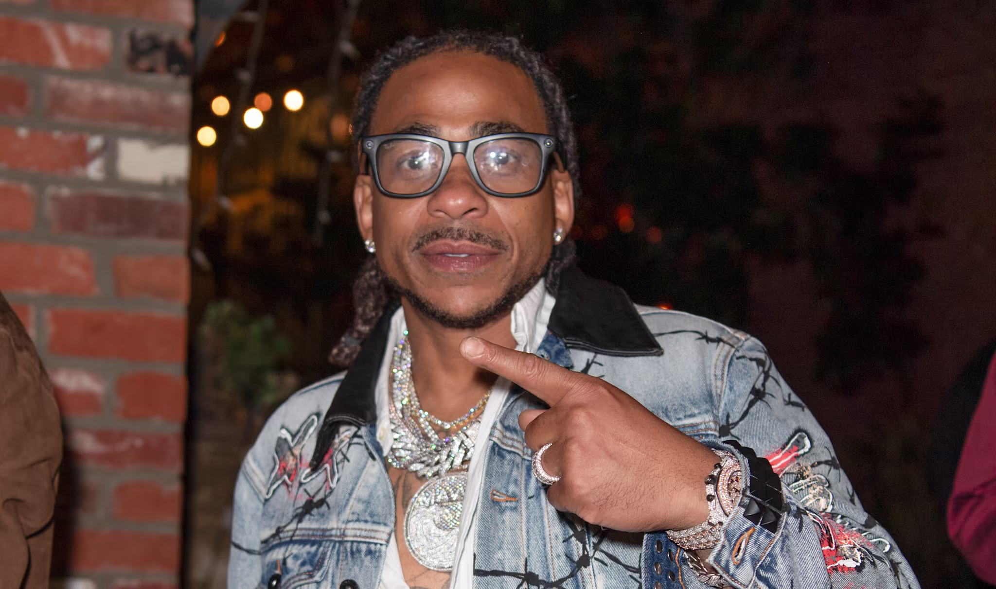 Max B wearing glasses, a denim jacket with barbed wire design, and a white shirt, pointing at himself, with a background of brick walls.