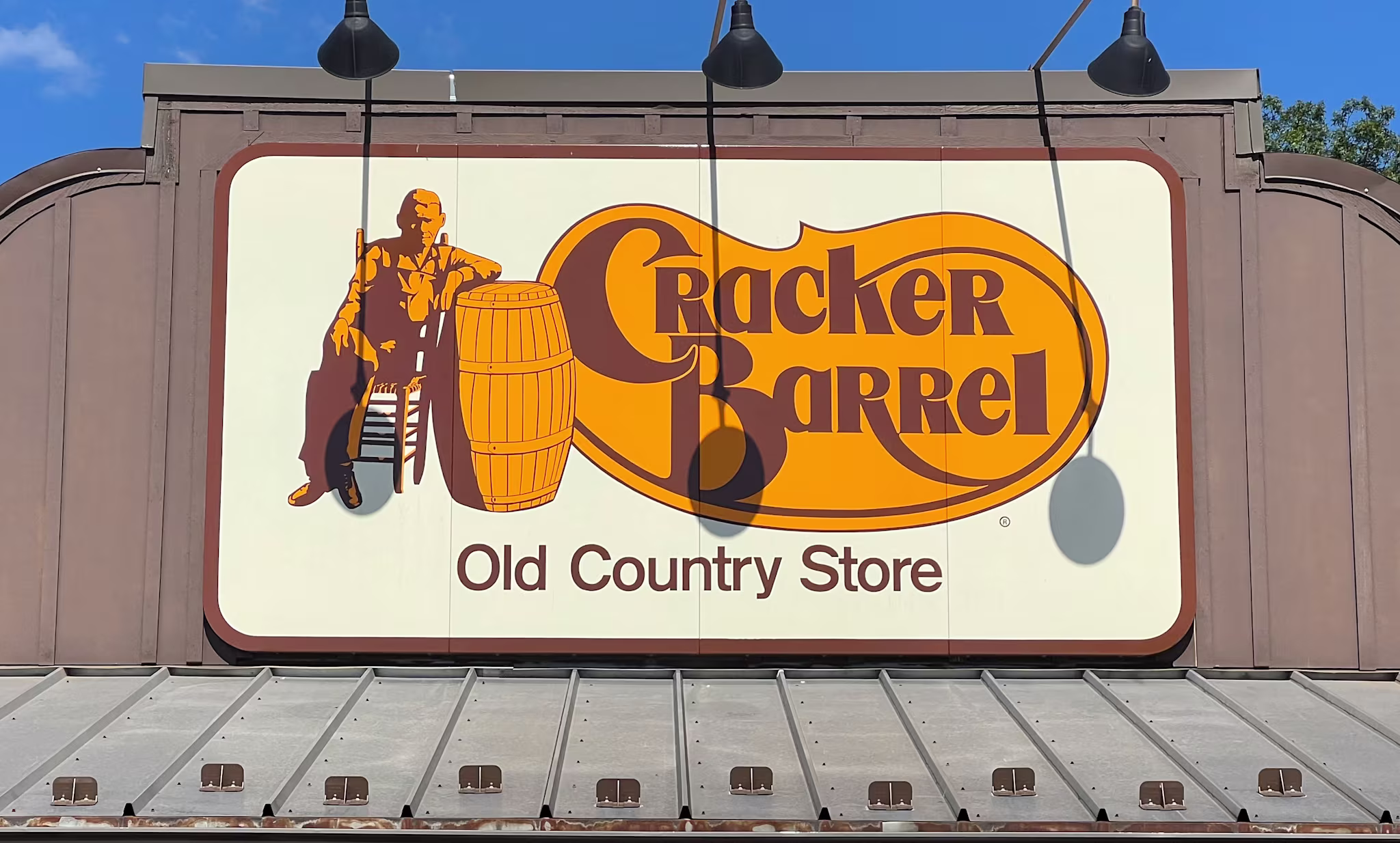 Sign for Cracker Barrel Old Country Store featuring a man sitting on a chair next to a barrel, with a blue sky background.