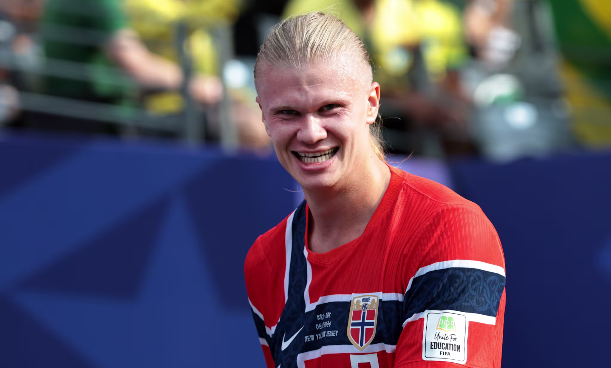 Erling Haaland in a red Norway jersey smiles on the field, with fans in the background.