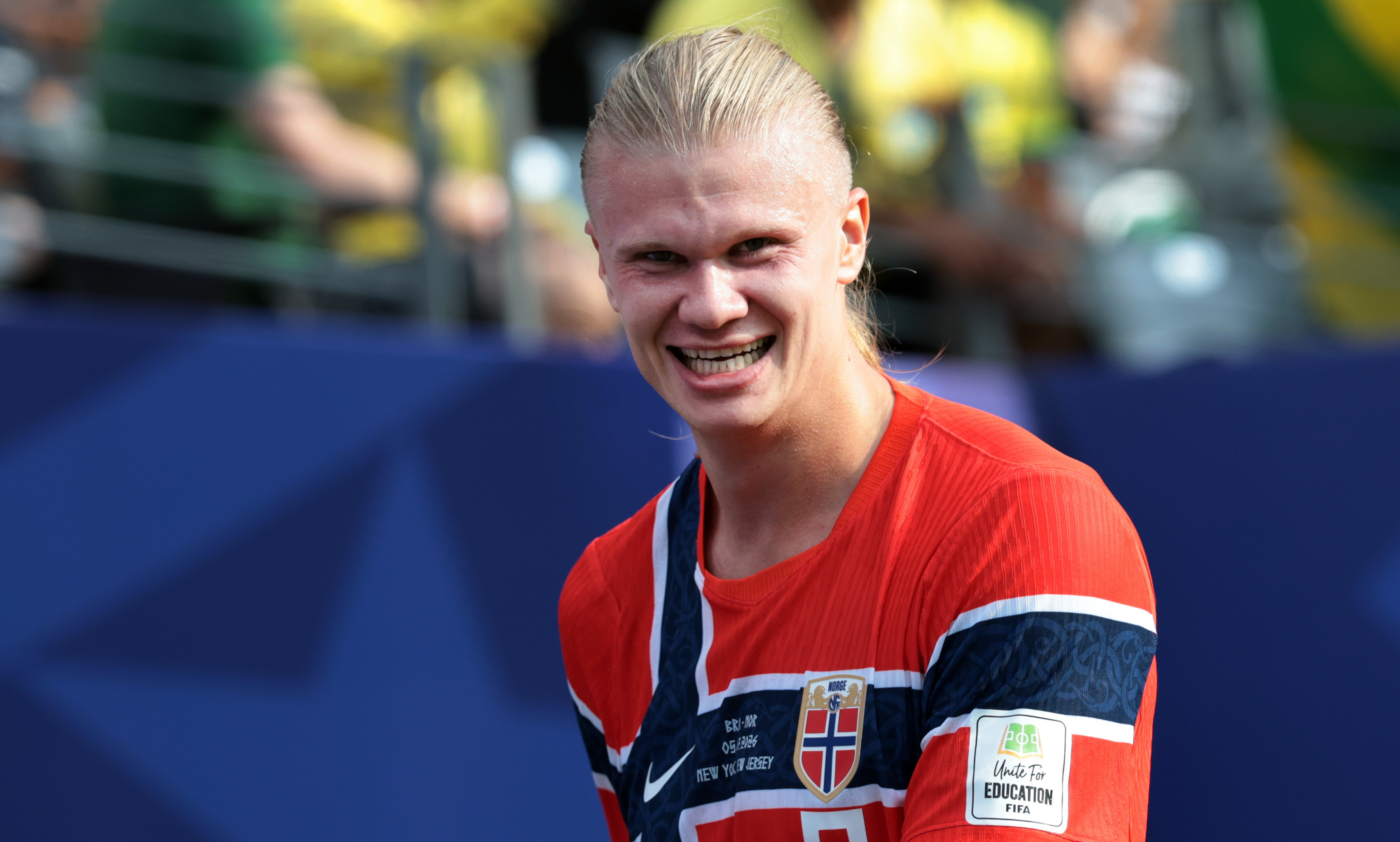 Erling Haaland in a red Norway jersey smiles on the field, with fans in the background.