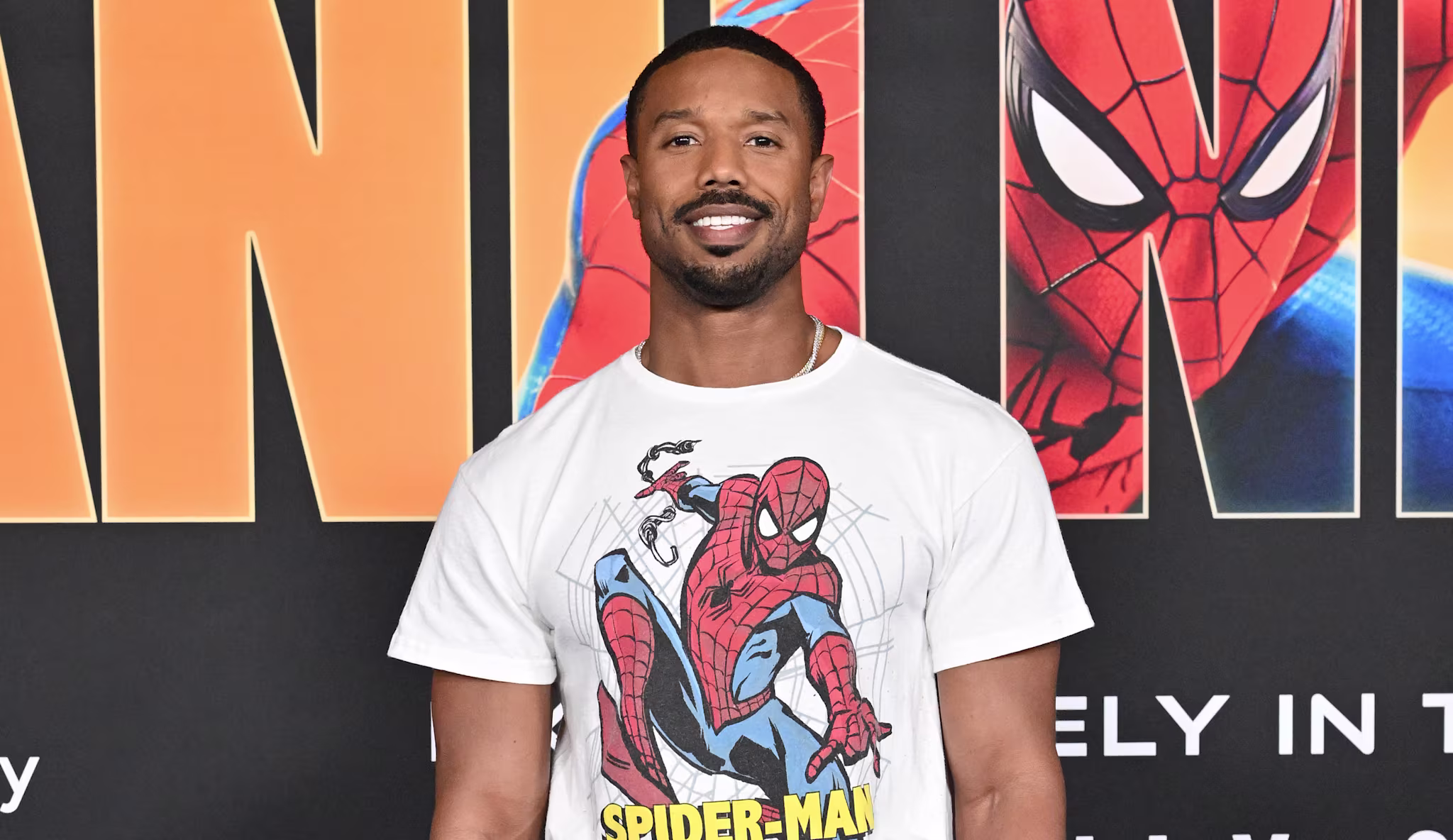 Michael B. Jordan at a Spider-Man event, wearing a Spider-Man t-shirt and black pants, standing on a blue carpet.