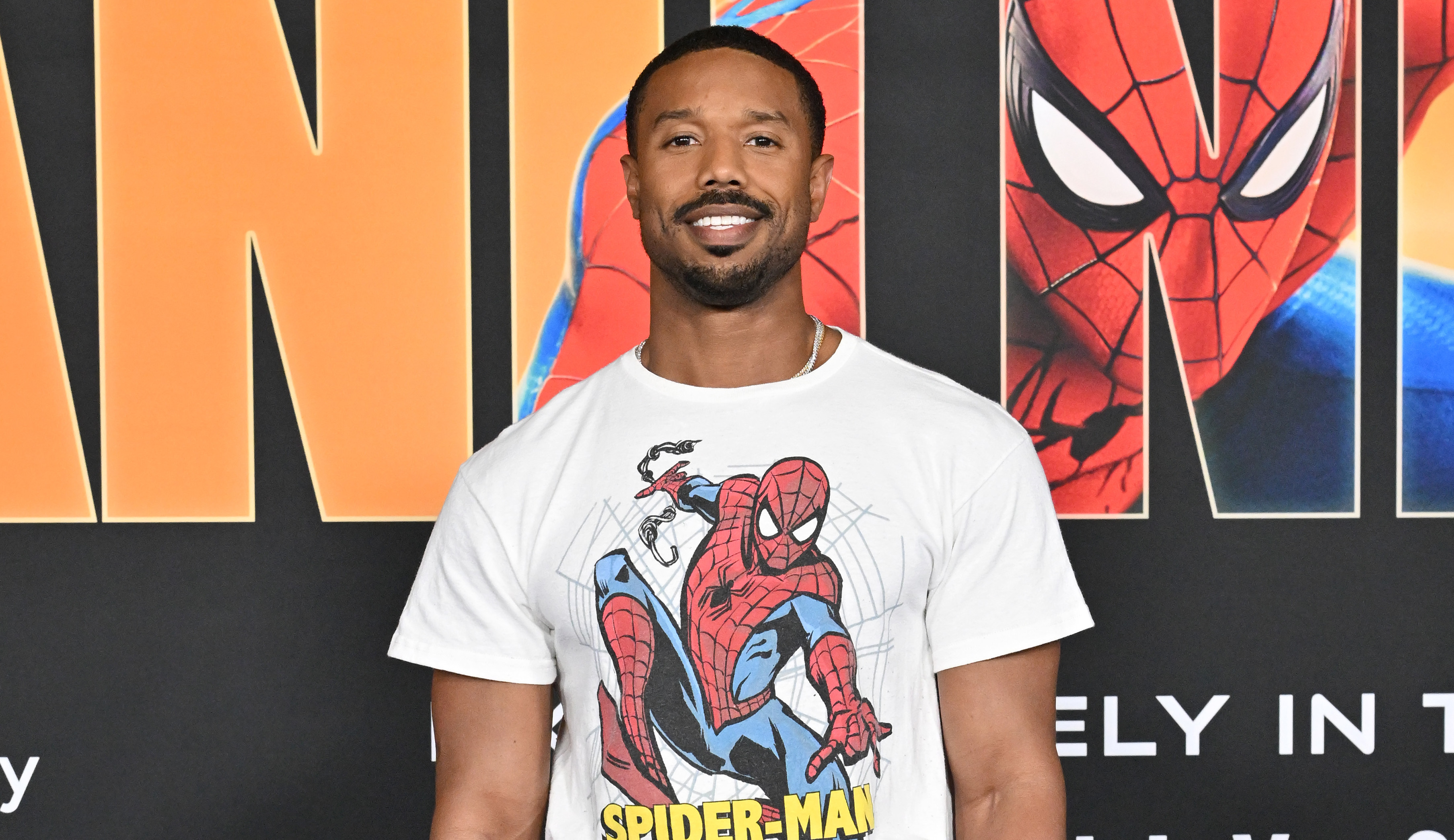 Michael B. Jordan at a Spider-Man event, wearing a Spider-Man t-shirt and black pants, standing on a blue carpet.