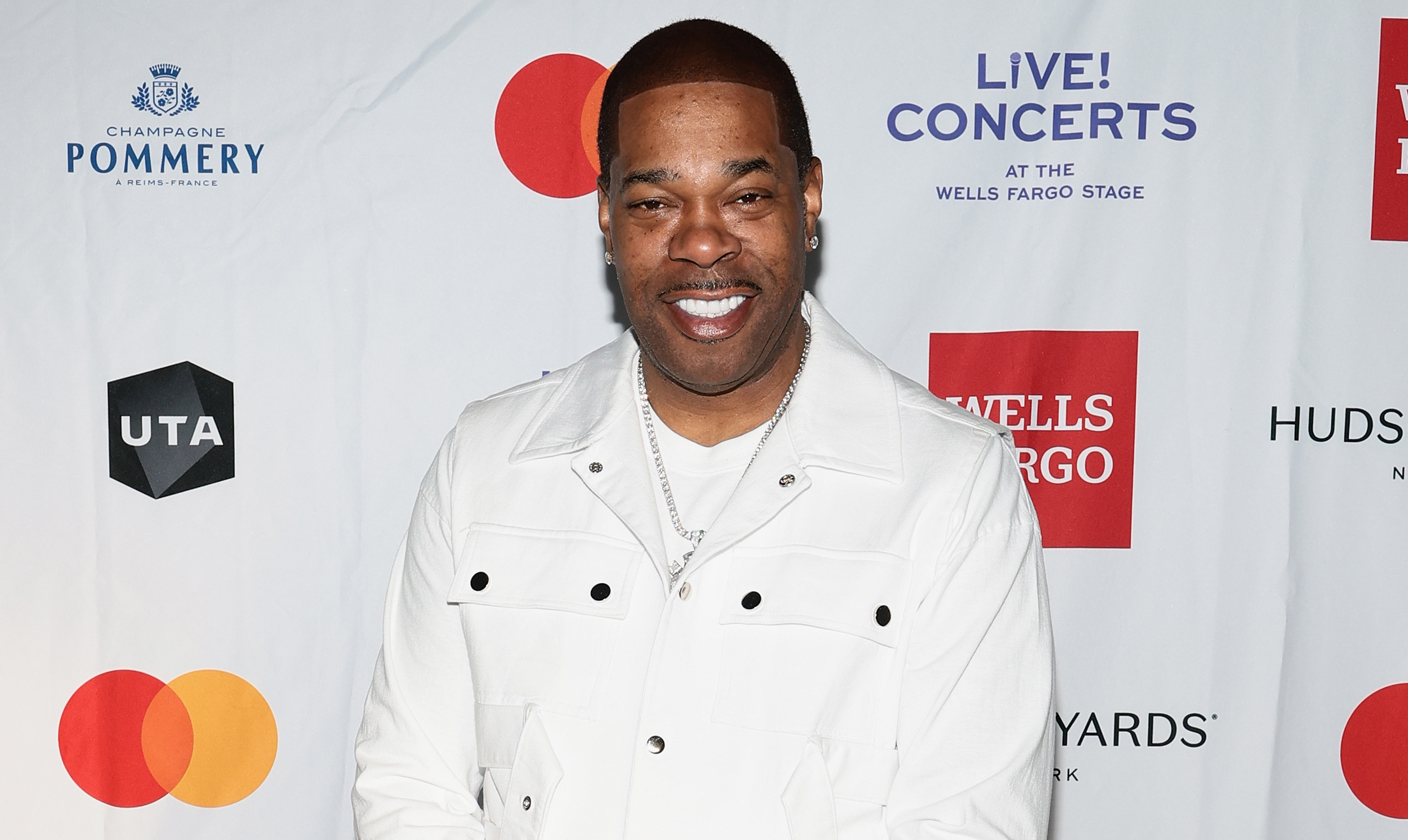 Busta Rhymes in a white outfit stands smiling on a branded backdrop, featuring logos like Wells Fargo and Mastercard.
