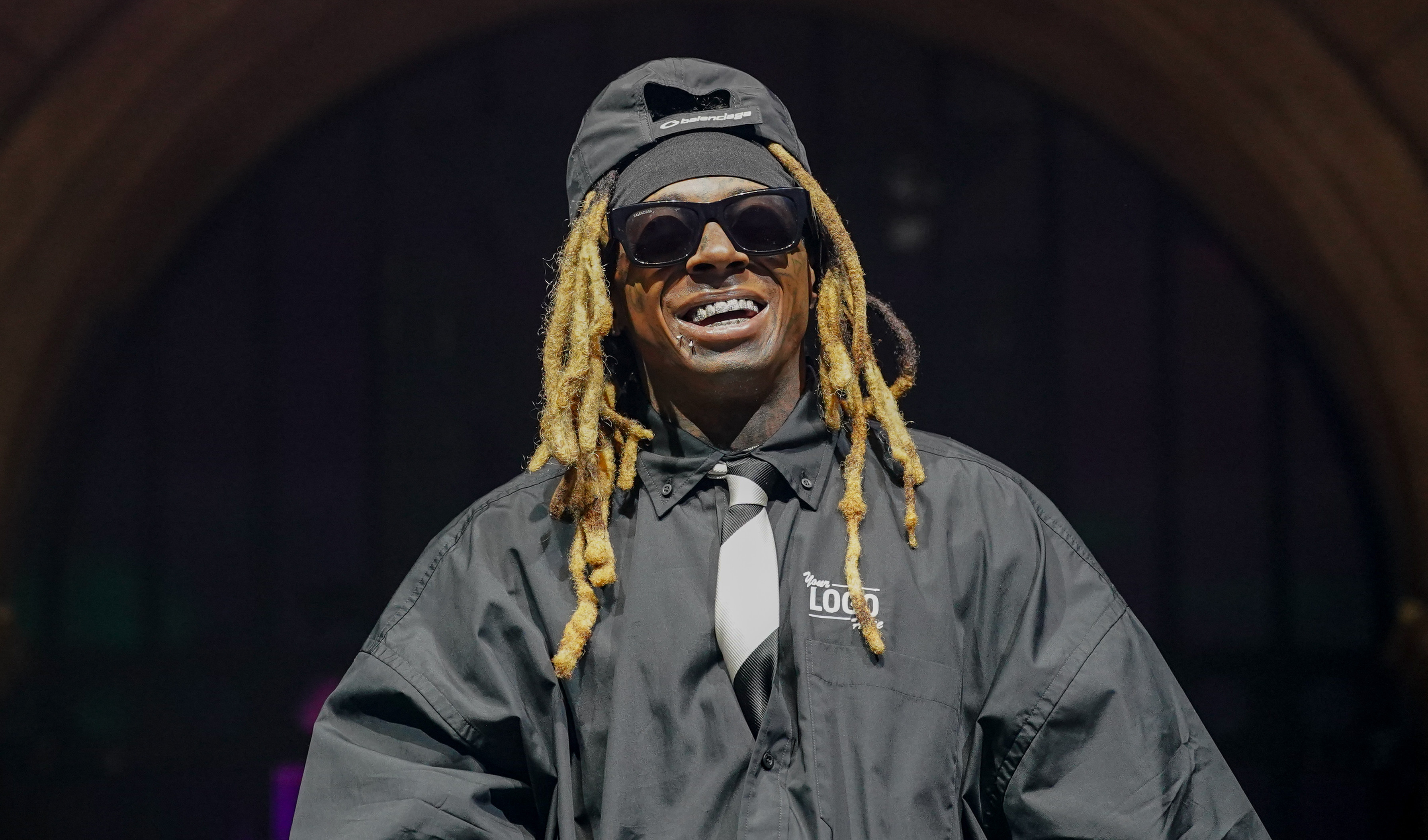 Lil Wayne performing on stage, wearing sunglasses and a black jacket, with "Tha Carter" sign in the background.