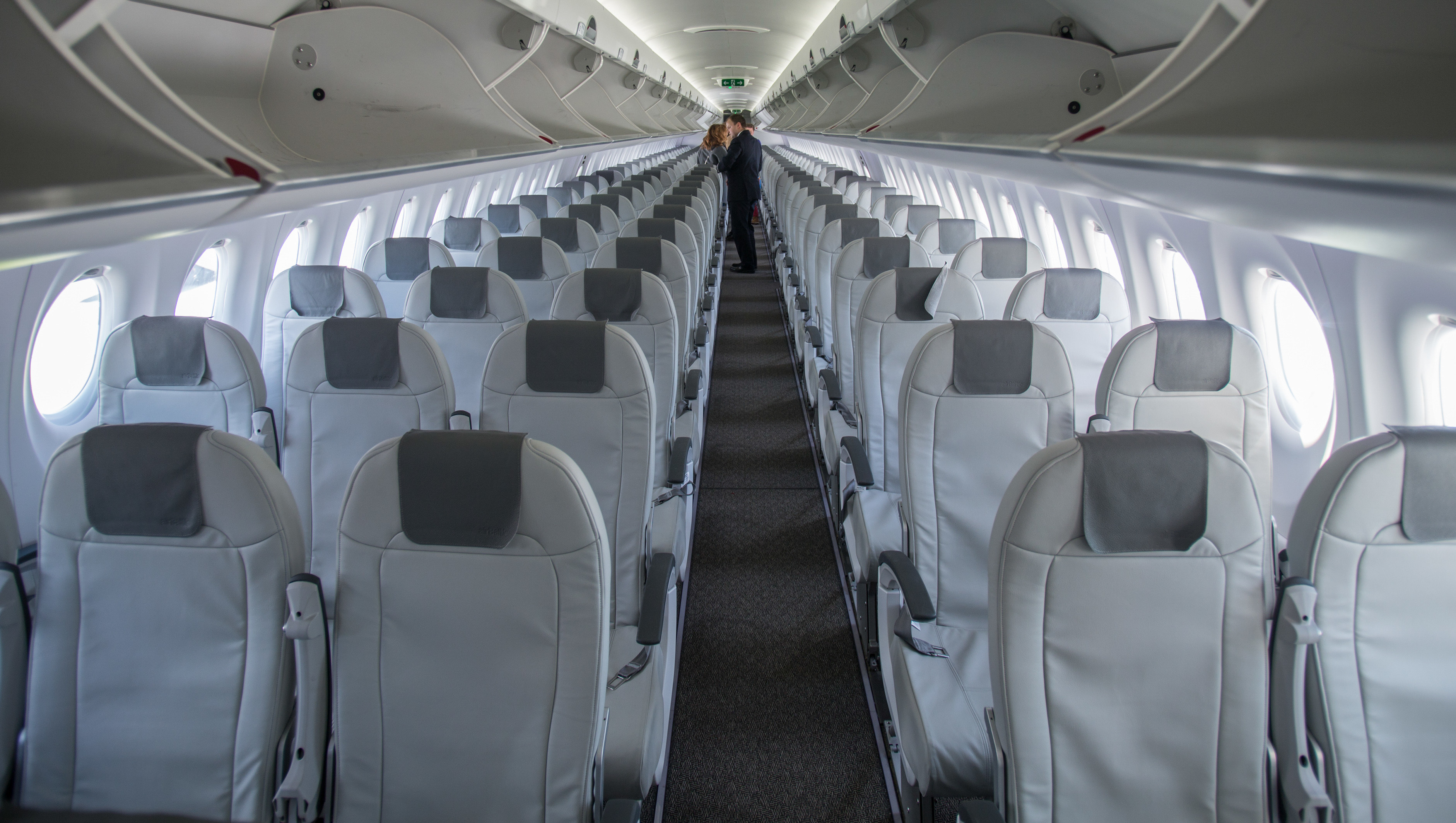 Empty airplane cabin with rows of gray seats, overhead bins open, and a person standing at the far end.